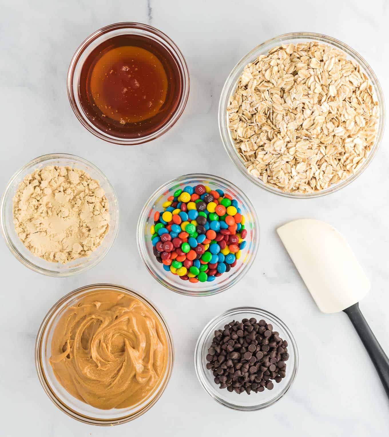 the ingredients in separate clear bowls - peanut butter, honey, protein powder, rolled oats, mini chocolate chips, and mini m&m's