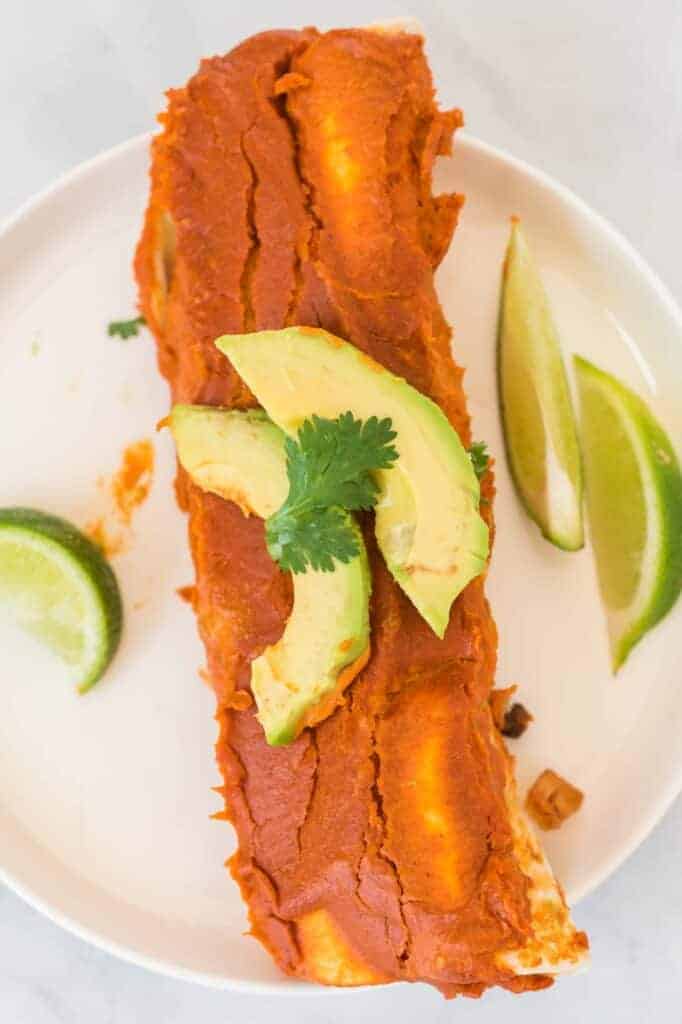 two enchiladas on a white plate topped with avocado and cilantro, with limes on the side
