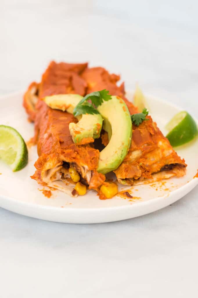 two enchiladas on a white plate topped with avocado and cilantro, with limes on the side