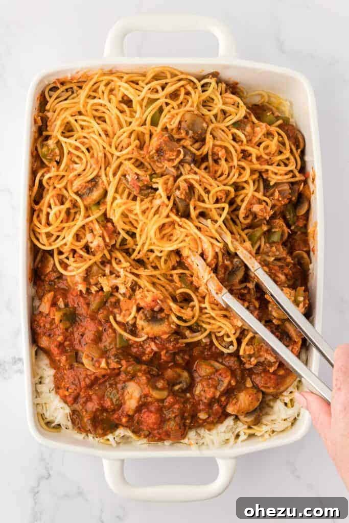 A pair of tongs gently mixing the spaghetti and sauce together in the casserole dish, ensuring even coating.