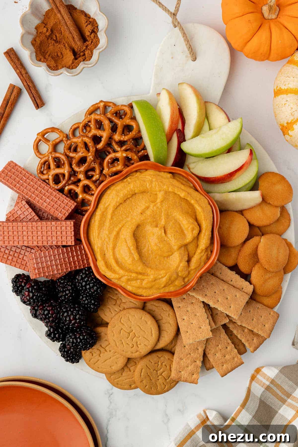 Pumpkin dip artfully arranged on a rustic board, surrounded by an assortment of crackers, crisp apple slices, savory pretzels, and vibrant fresh fruit