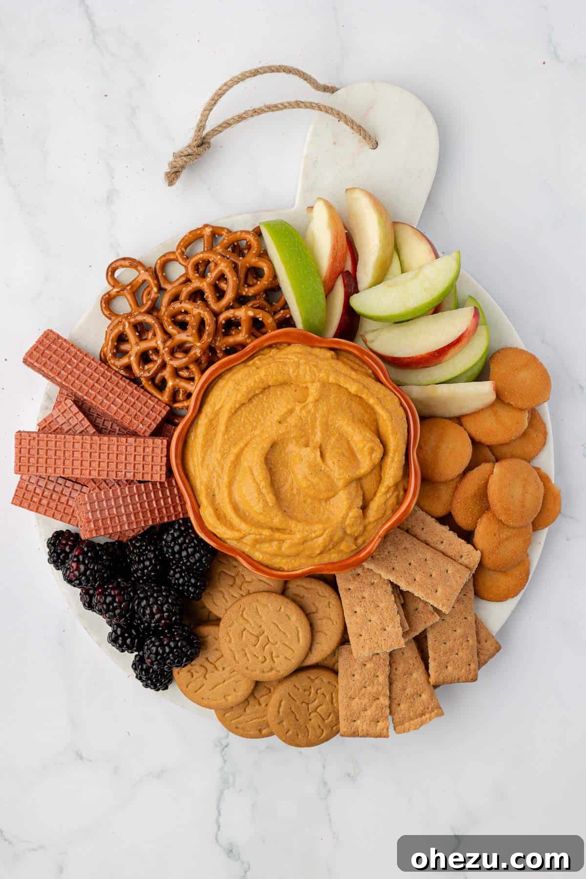 Beautifully arranged pumpkin dip on a wooden board, surrounded by an assortment of crackers, fresh apple slices, crunchy pretzels, and other appealing fruit