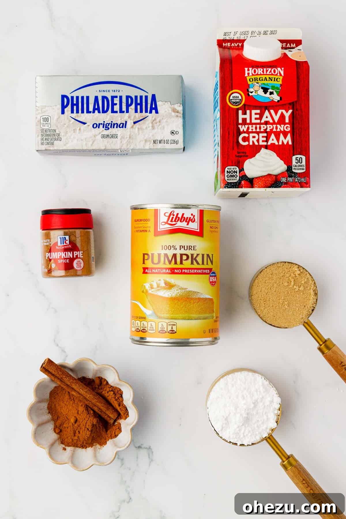 Individual ingredients for pumpkin dip artfully arranged: a block of cream cheese, a carton of heavy cream, a can of pumpkin puree, a bowl of brown sugar, a spoon of pumpkin pie spice, a bowl of powdered sugar, and a small pile of cinnamon