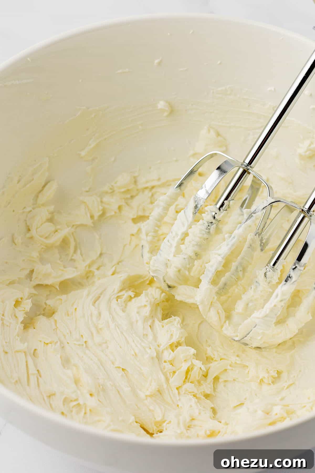Softened cream cheese being whipped to a smooth consistency in a clear mixing bowl