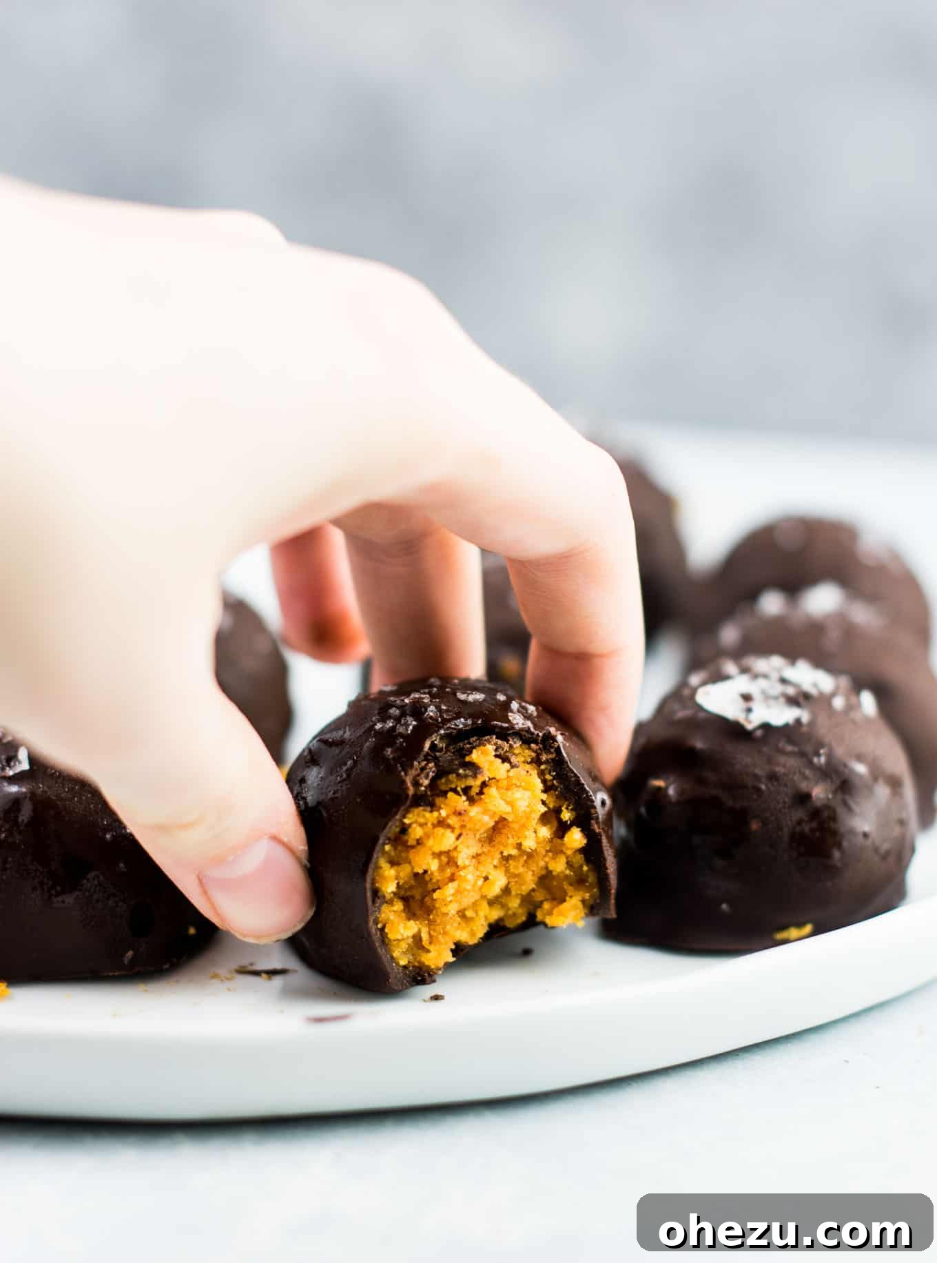 A hand reaching for a dark chocolate pumpkin truffle from a plate, highlighting its tempting appearance.