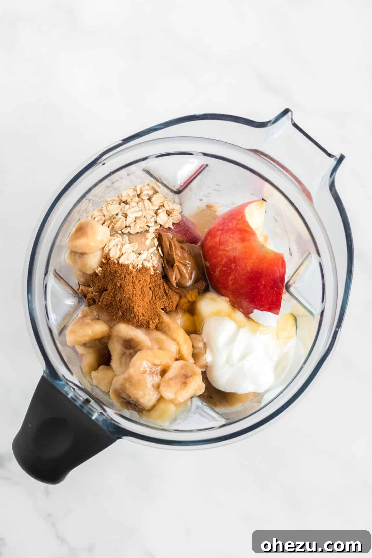 All the delicious apple pie smoothie ingredients, including apples, frozen bananas, Greek yogurt, cinnamon, honey, oats, and almond butter, placed inside a blender.