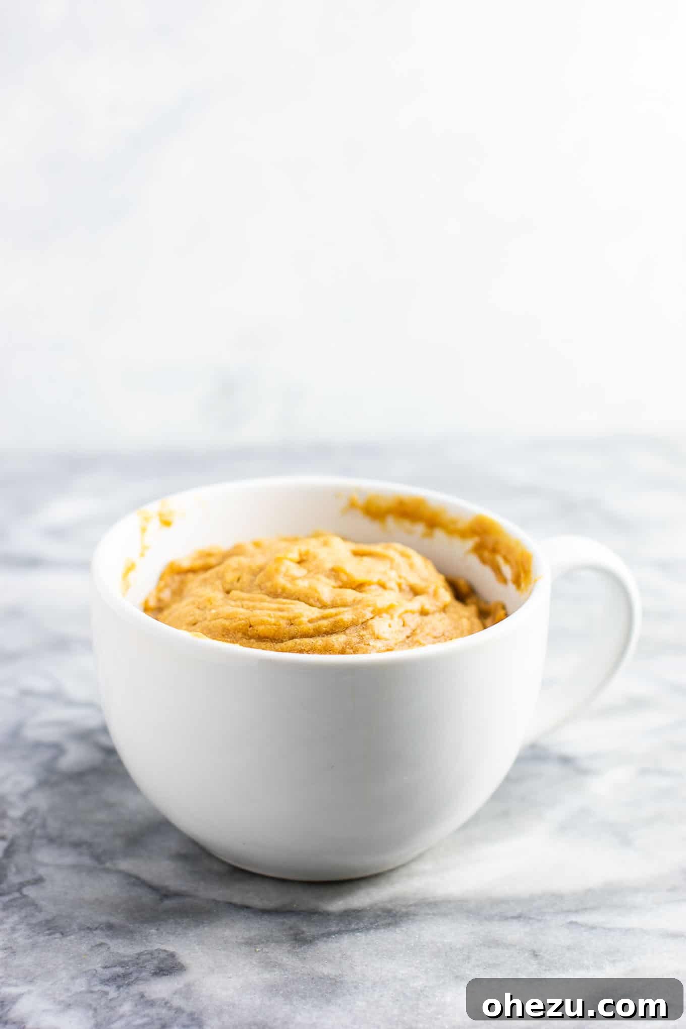 Step-by-step guide showing melted peanut butter in a mug, ready for mixing other gluten-free, dairy-free, refined sugar-free ingredients to make a microwave peanut butter cookie.