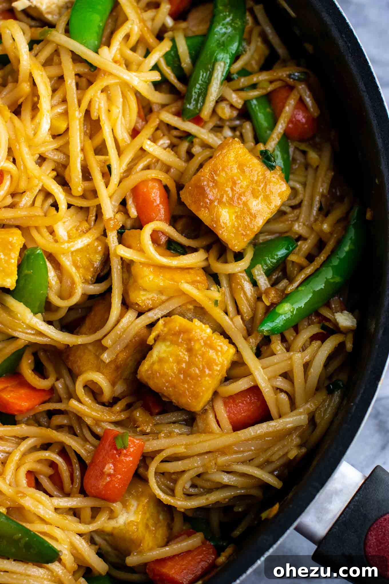 A large skillet brimming with colorful vegan stir fry noodles, tofu, sugar snap peas, and carrots.