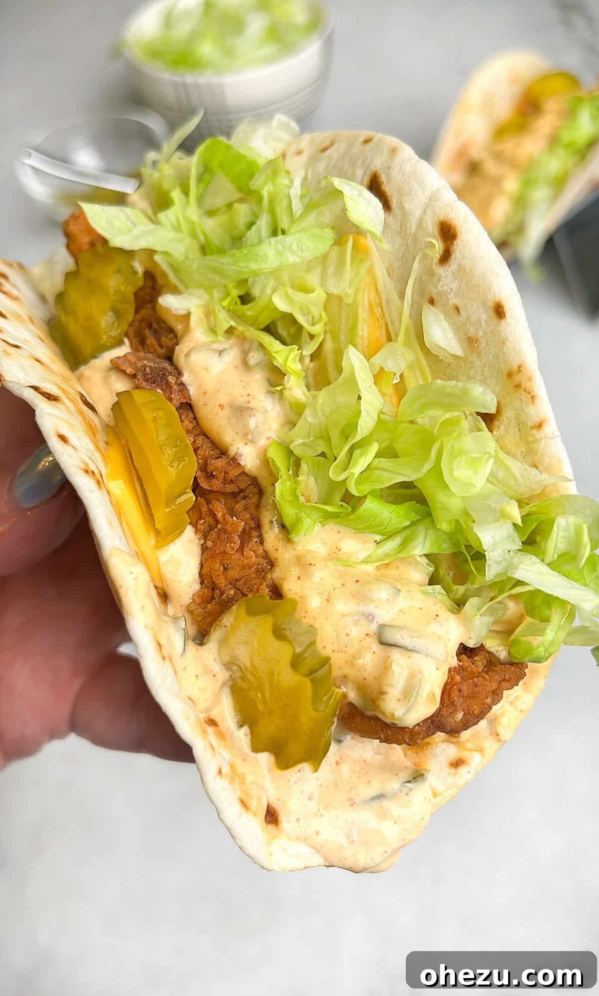 Ultimate Chicken Big Mac Taco 2 A crispy chicken Big Mac taco being held in a hand, showcasing its vibrant layers of sauce, lettuce, pickles, and golden-fried chicken against a soft flour tortilla.