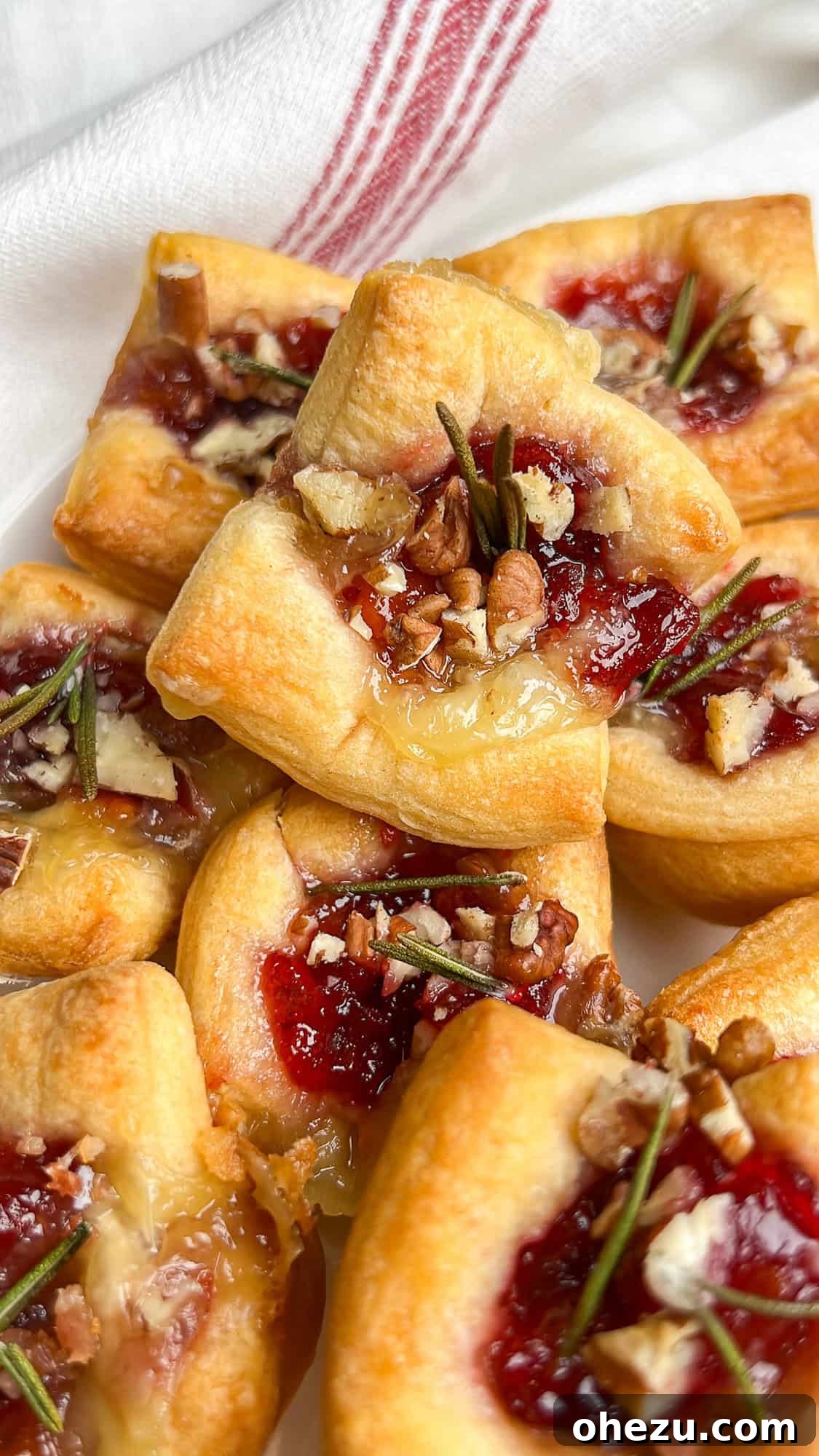 Flaky Cranberry Brie Bake 2 Beautifully baked brie and cranberry puff pastry bites, garnished with rosemary and pecans, stacked artfully on a plate, ready to be served as a festive holiday appetizer.