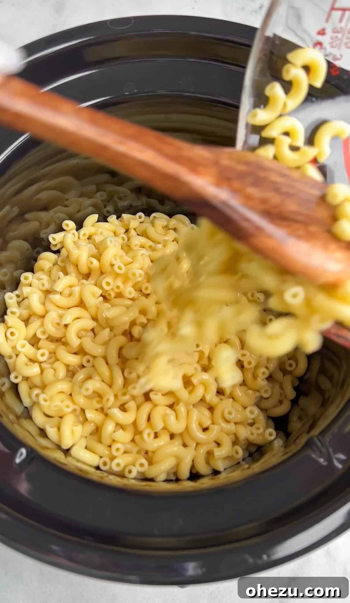Pouring partially cooked elbow macaroni, perfectly al dente, into the ceramic insert of a slow cooker.