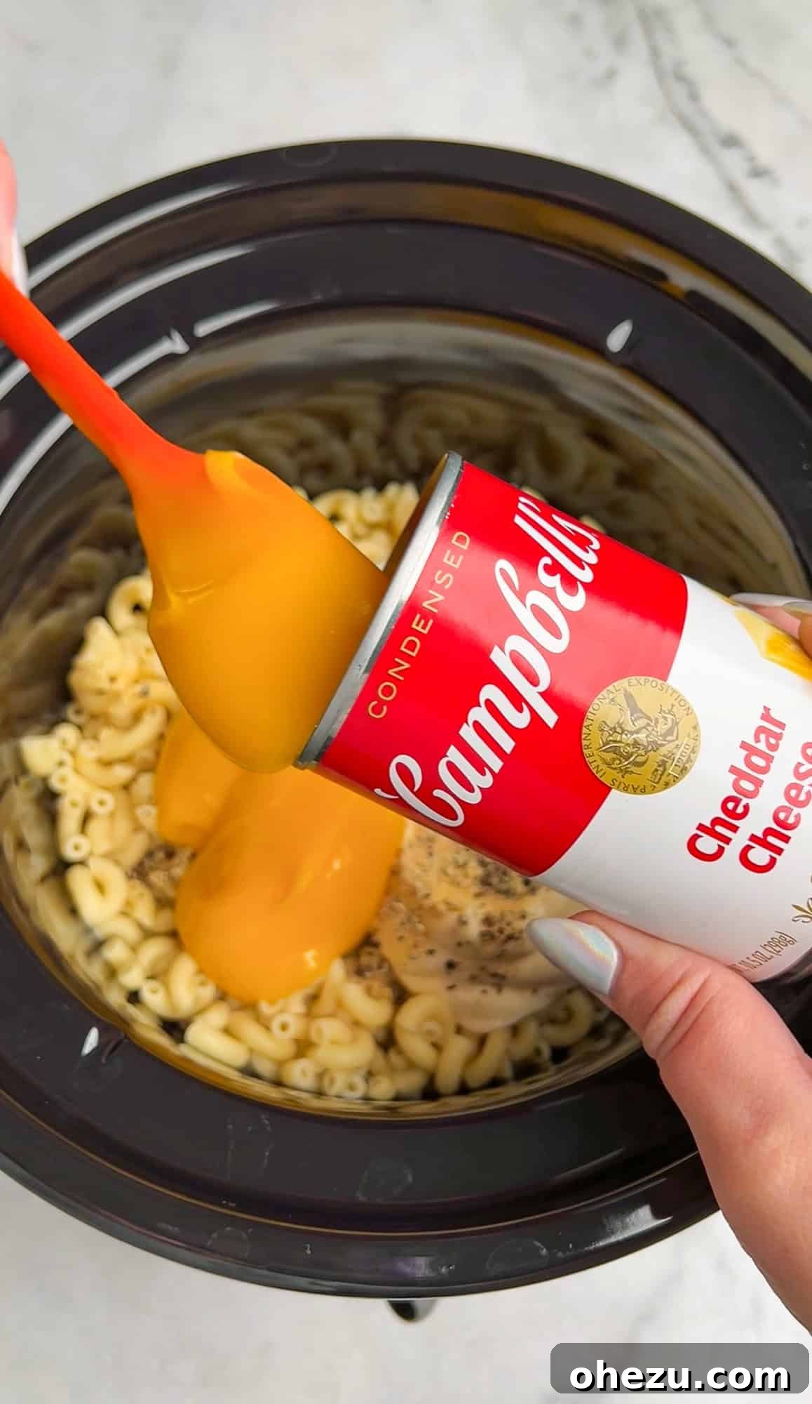 A hand spoons a can of condensed cheddar cheese soup over the par-cooked macaroni in the slow cooker, alongside other ingredients.