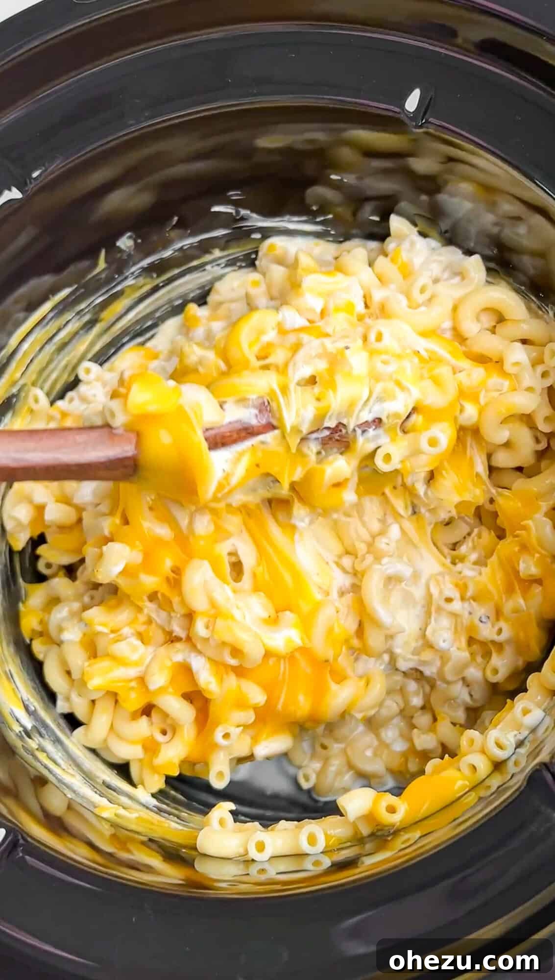 A wooden spoon actively stirring the macaroni, condensed soup, sour cream, mayonnaise, seasonings, and cheese within the slow cooker, creating a consistent mixture.