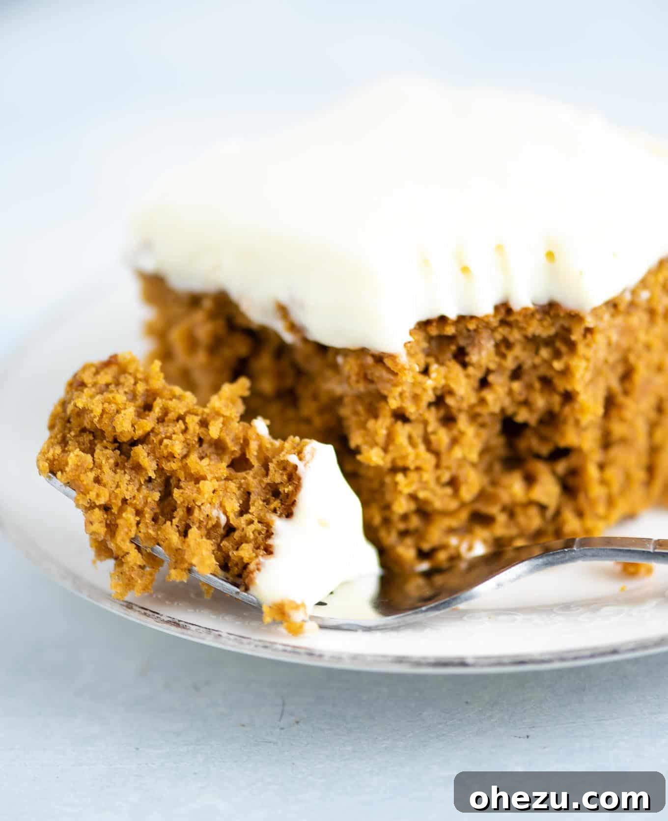 A close-up image of a fork taking a bite from a slice of pumpkin cake, showcasing the fluffy cream cheese frosting and the moist, orange cake layers.