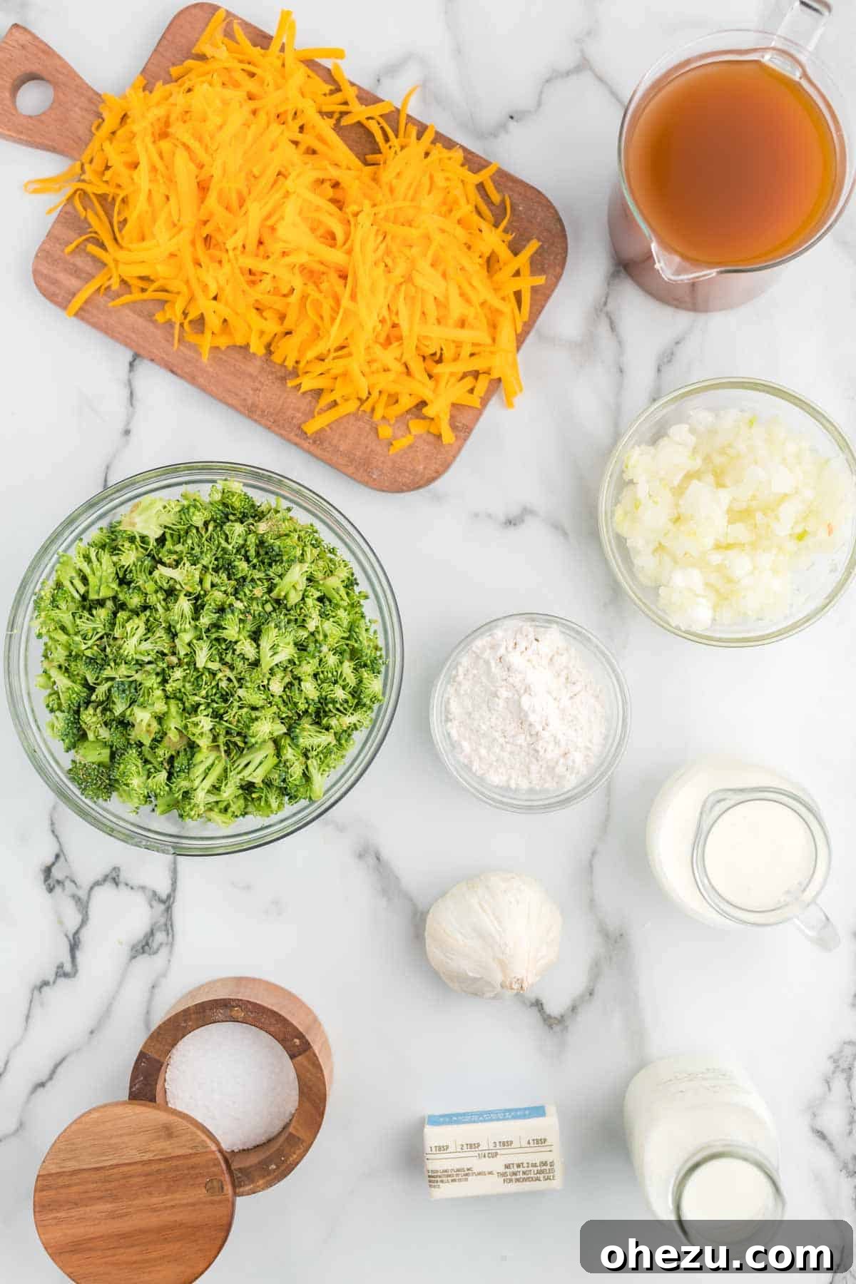 Fresh ingredients laid out beautifully for making homemade broccoli cheese soup: butter, flour, onion, garlic, whole milk, vegetable broth, heavy cream, broccoli florets, and a block of sharp cheddar cheese on a wooden board.