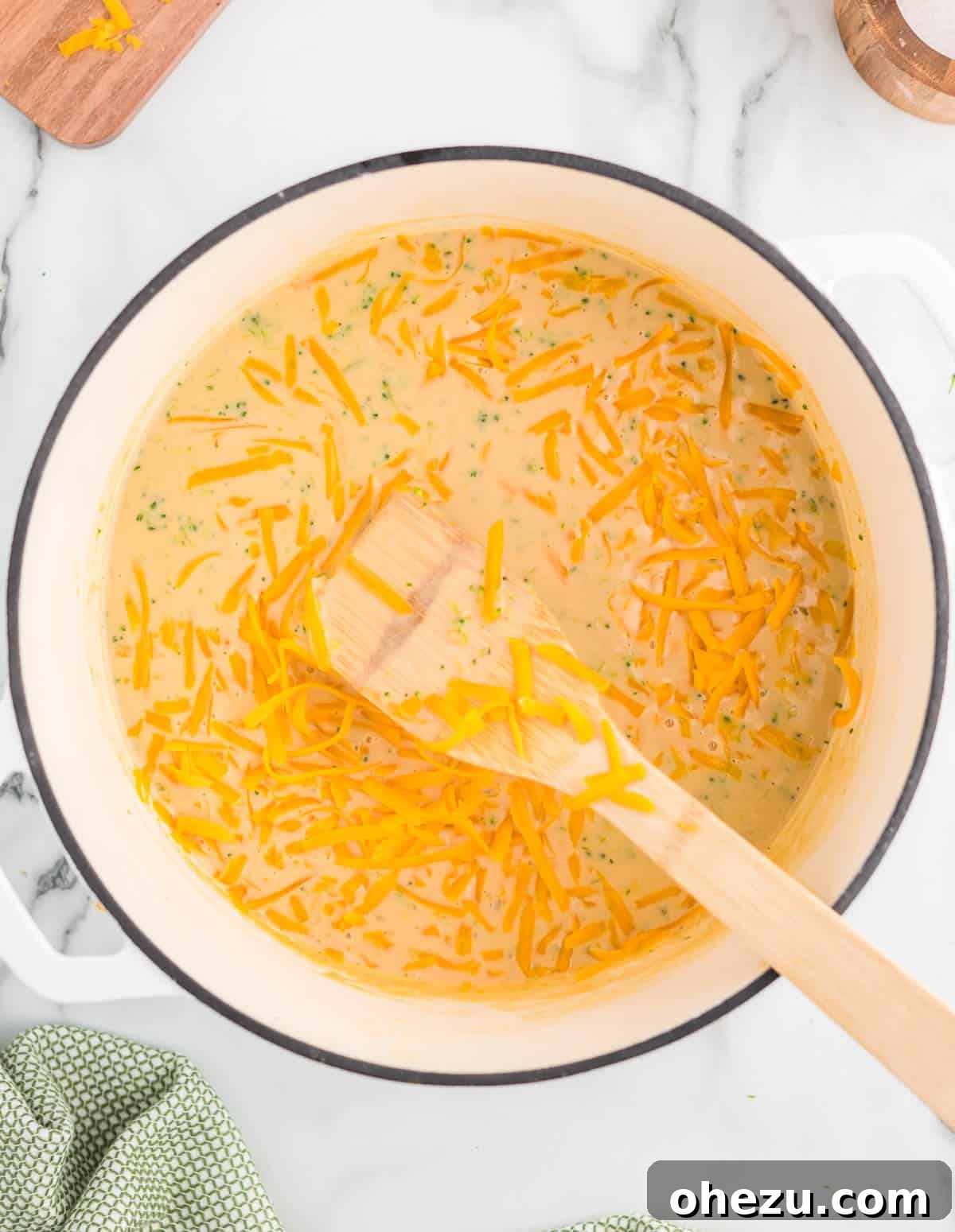 A hand adding freshly shredded sharp cheddar cheese into the simmering pot of broccoli cheese soup, stirring gently.