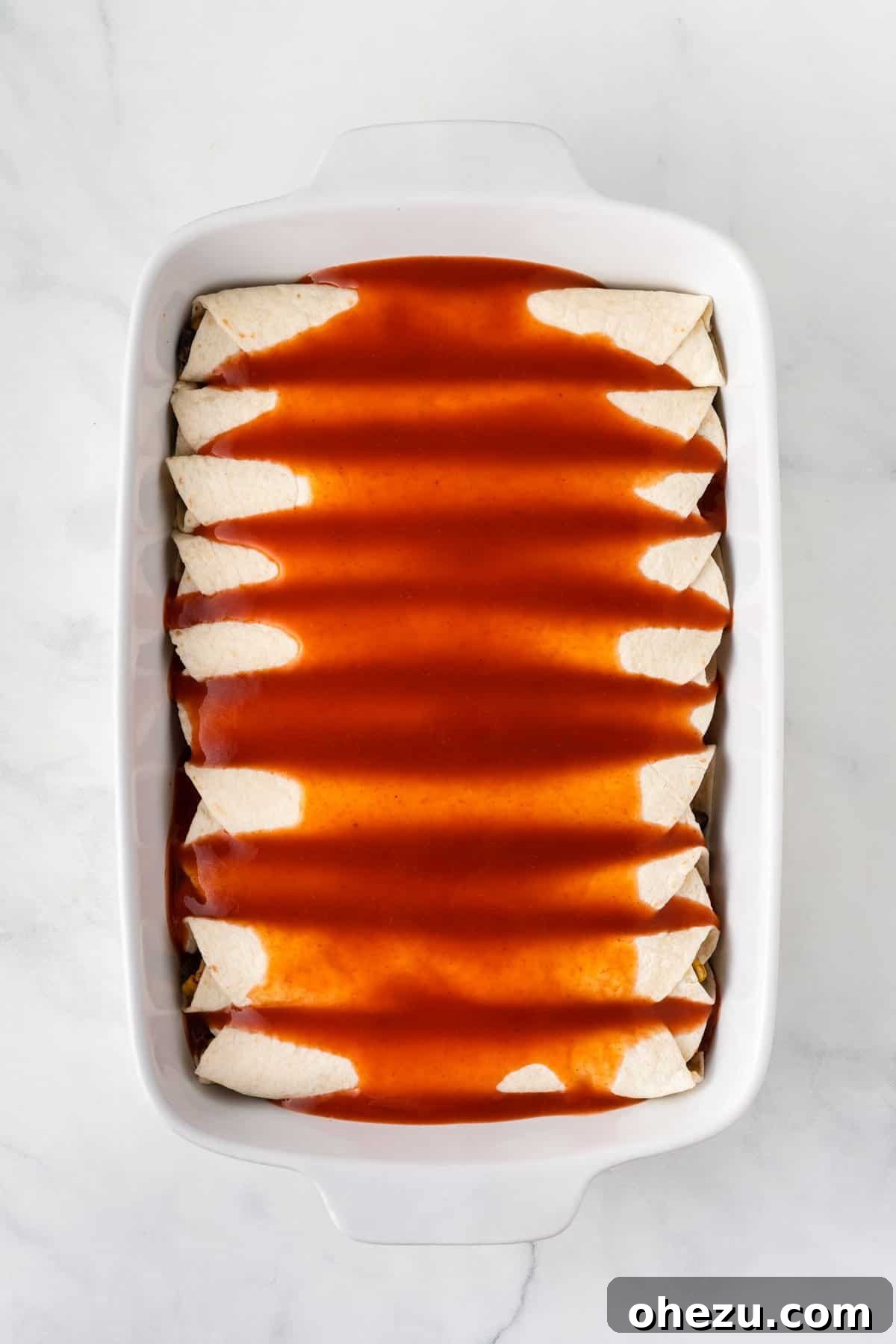 Carefully pouring rich red enchilada sauce over the assembled vegetarian enchiladas in the baking dish for even coverage.