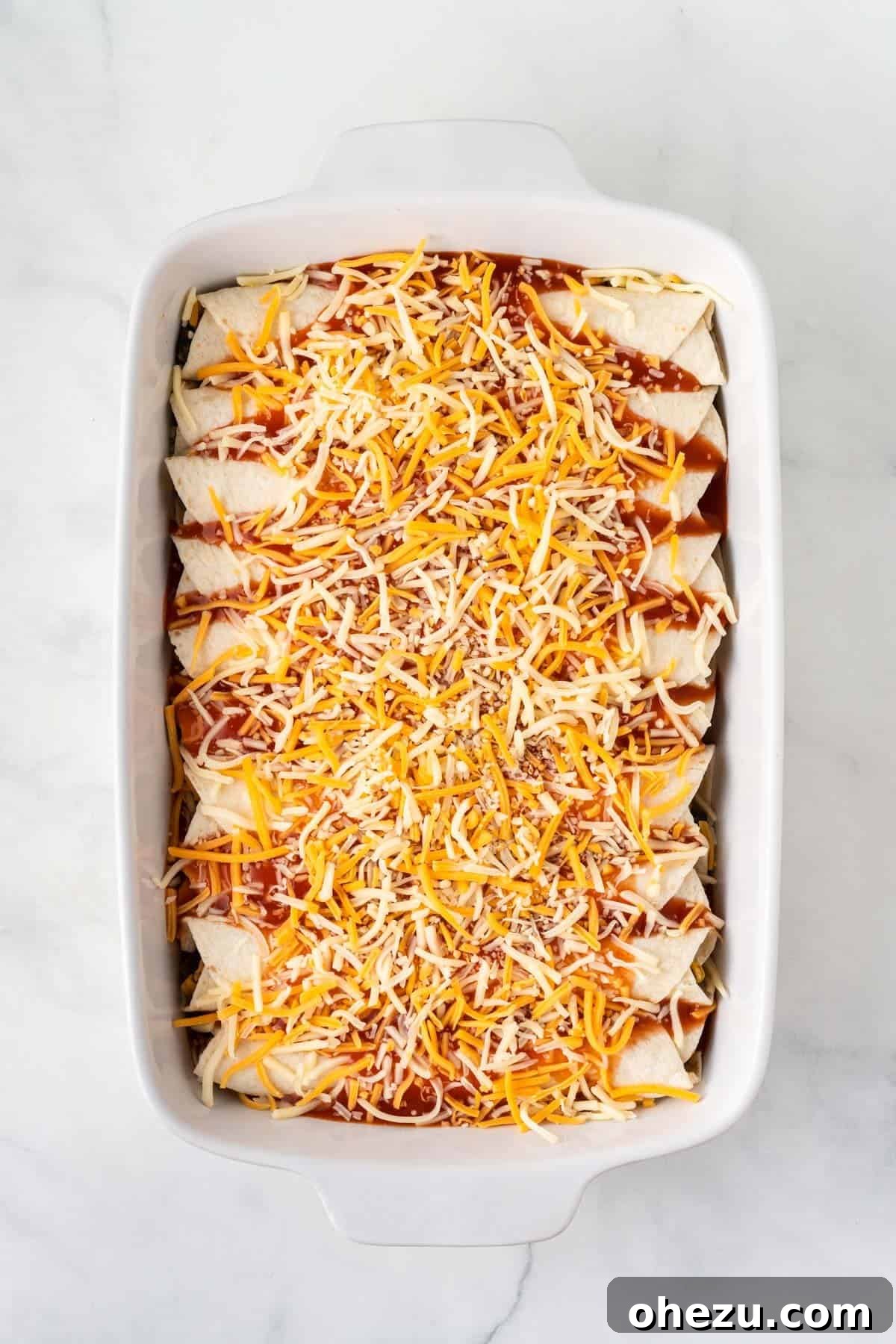 Generously sprinkling shredded Mexican cheese over the sauce-covered enchiladas, preparing them for baking.