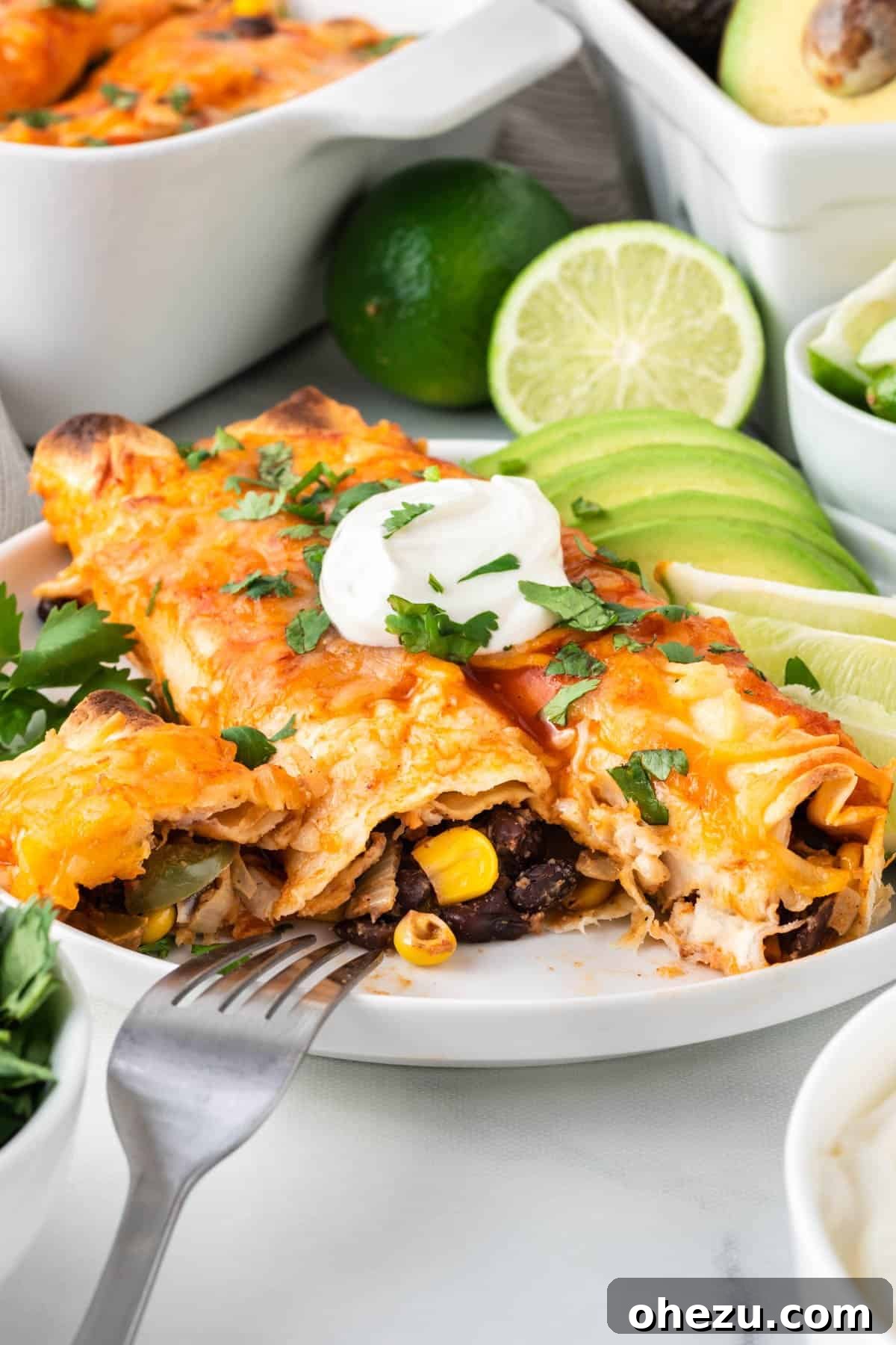 A serving of golden-baked vegetarian enchiladas on a white plate, elegantly garnished with cool sour cream, fresh lime wedges, and vibrant avocado slices.