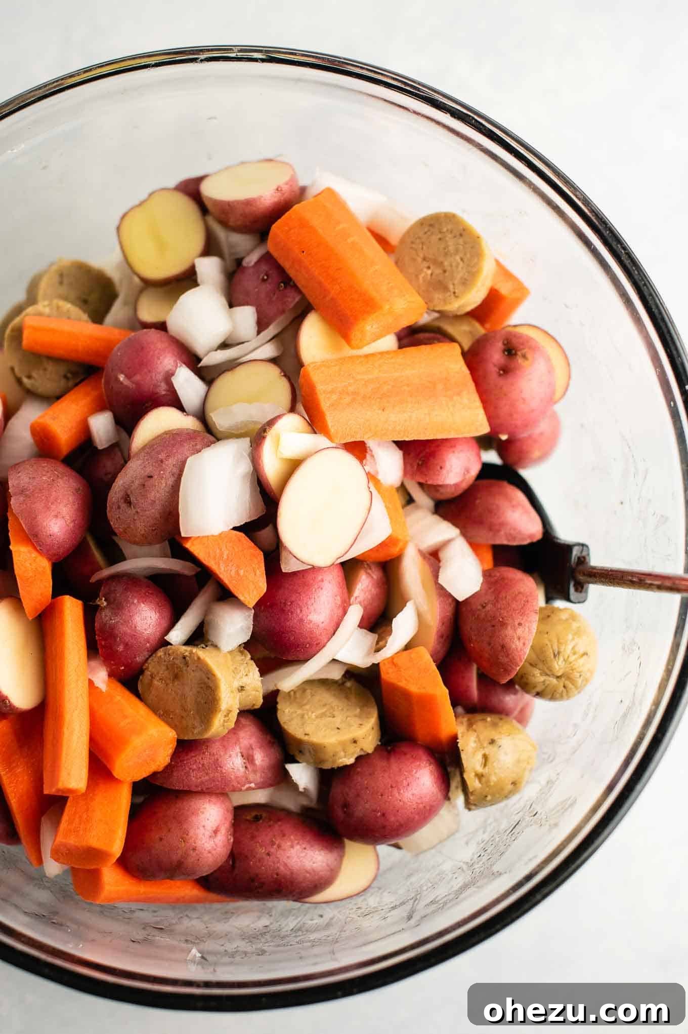 Sliced potatoes, carrots, onions, and vegetarian kielbasa from an overhead view, seasoned and coated in olive oil in a mixing bowl.