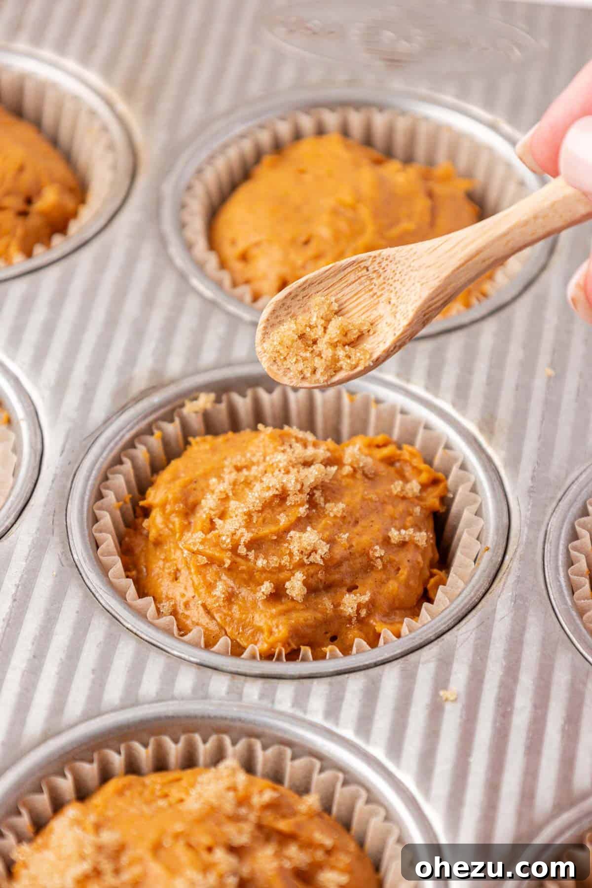 Hands sprinkling brown sugar over the tops of pumpkin muffin batter in a tin before baking