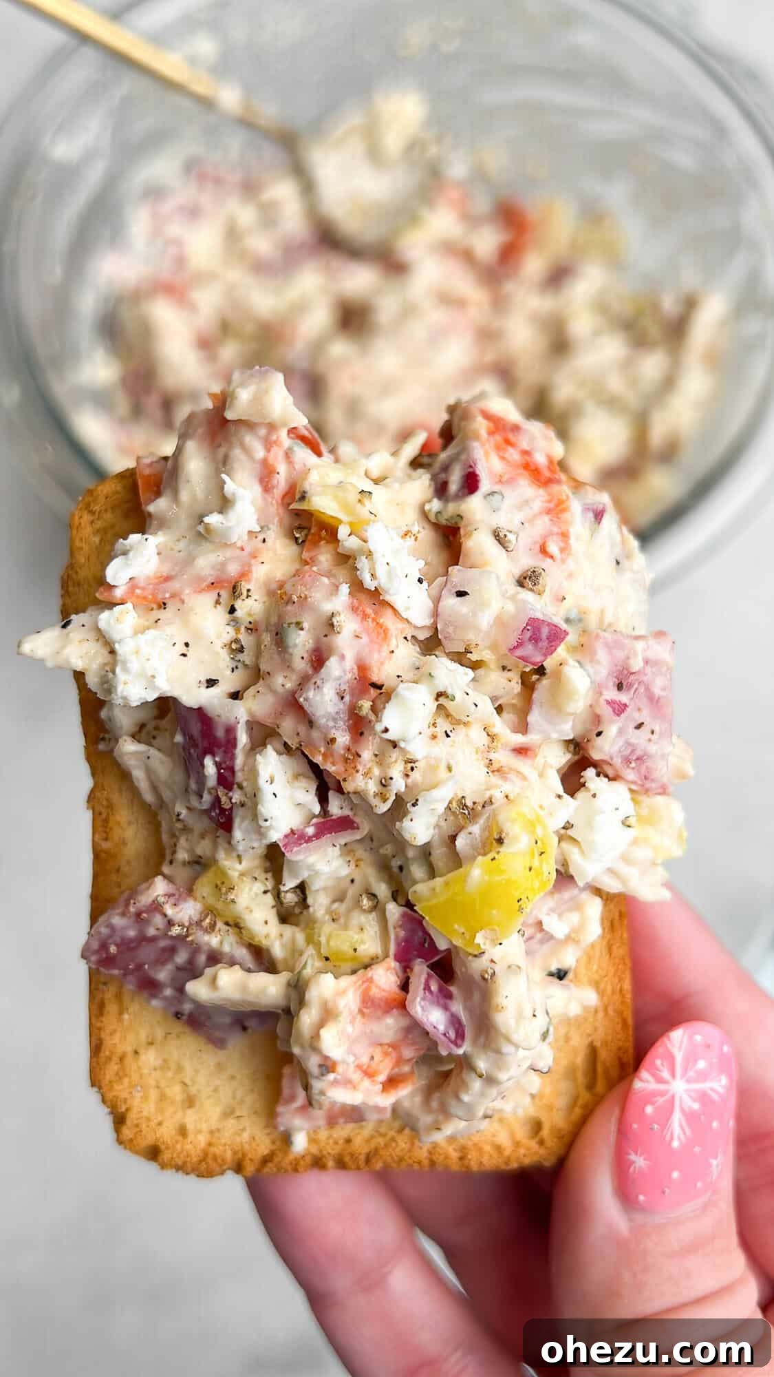 A hand holding a cracker topped with a generous scoop of Italian Grinder Chicken Salad, showcasing its vibrant ingredients.