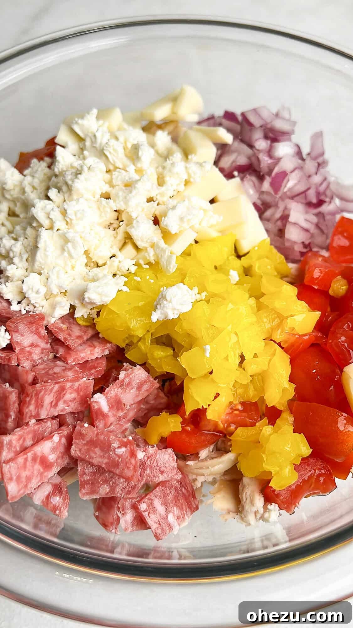 A bowl filled with all the colorful toppings for the Italian Grinder Chicken Salad, including deli meats, tomatoes, and banana peppers.