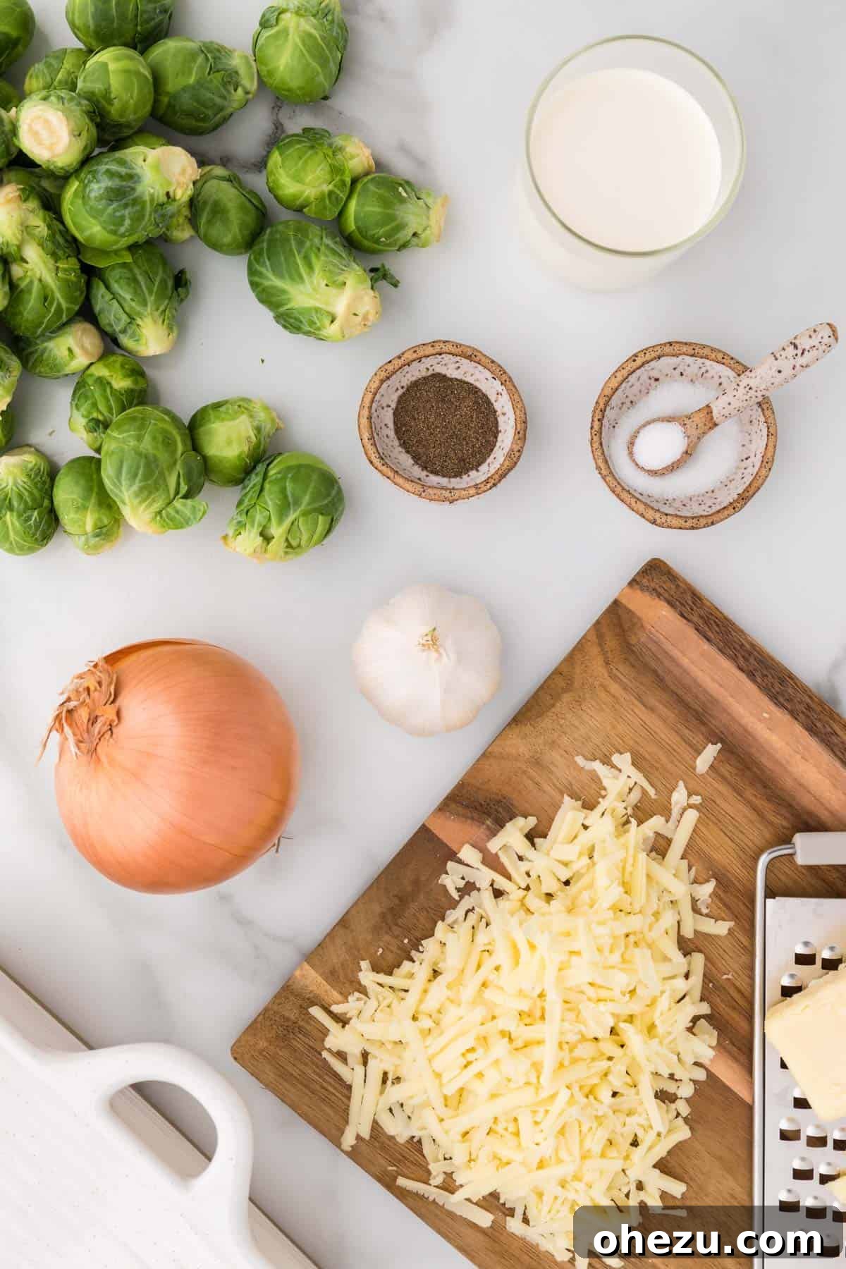 Ultimate Cheesy Brussels Sprouts 3 A collection of fresh ingredients laid out for cheesy Brussels sprouts gratin: green Brussels sprouts, yellow onion, garlic cloves, heavy cream, block of white cheddar cheese, salt, and pepper.