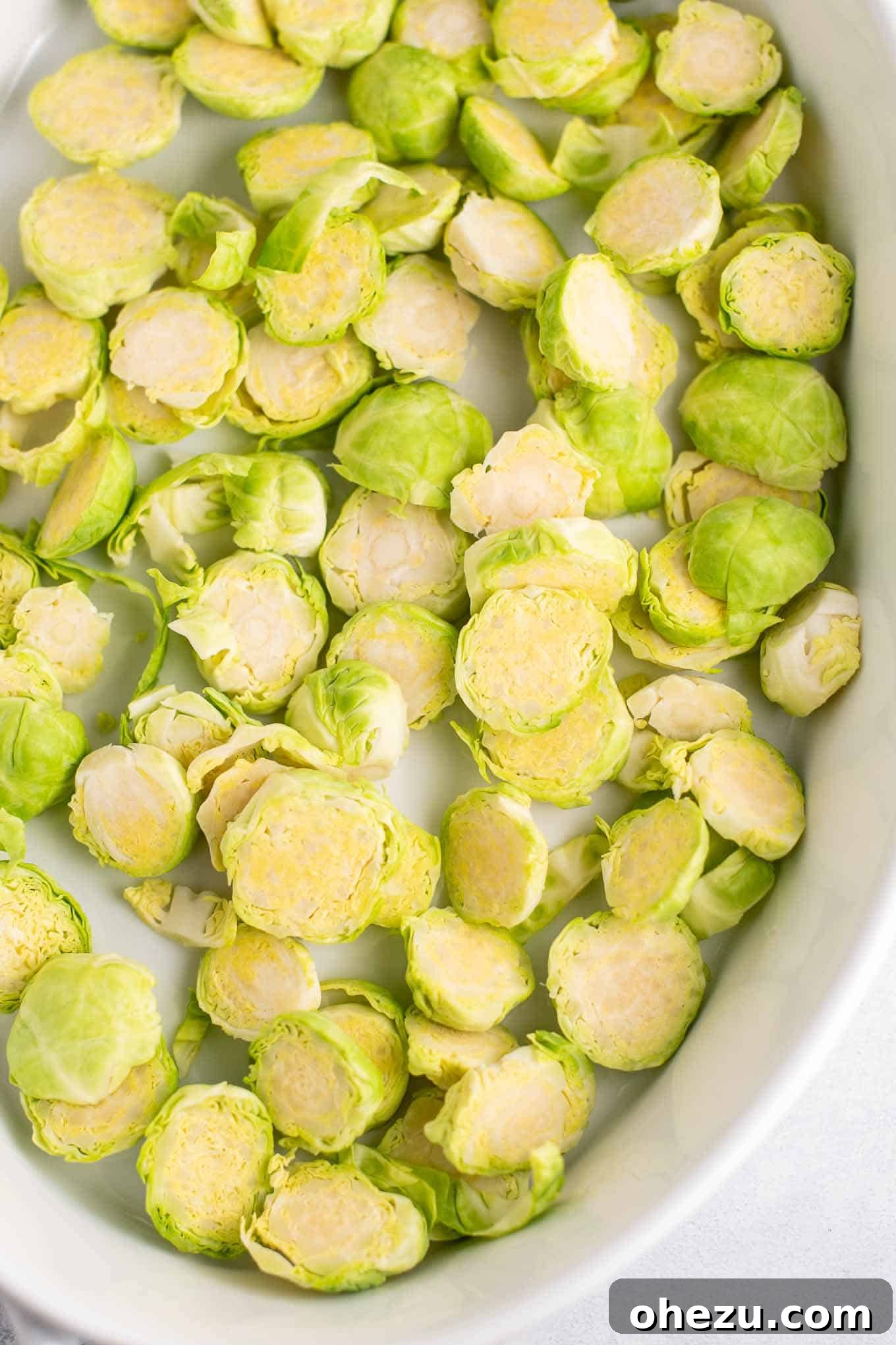 Ultimate Cheesy Brussels Sprouts 5 Thinly sliced raw Brussels sprouts arranged in a single layer within an oval white baking dish, prepared for the initial roasting.