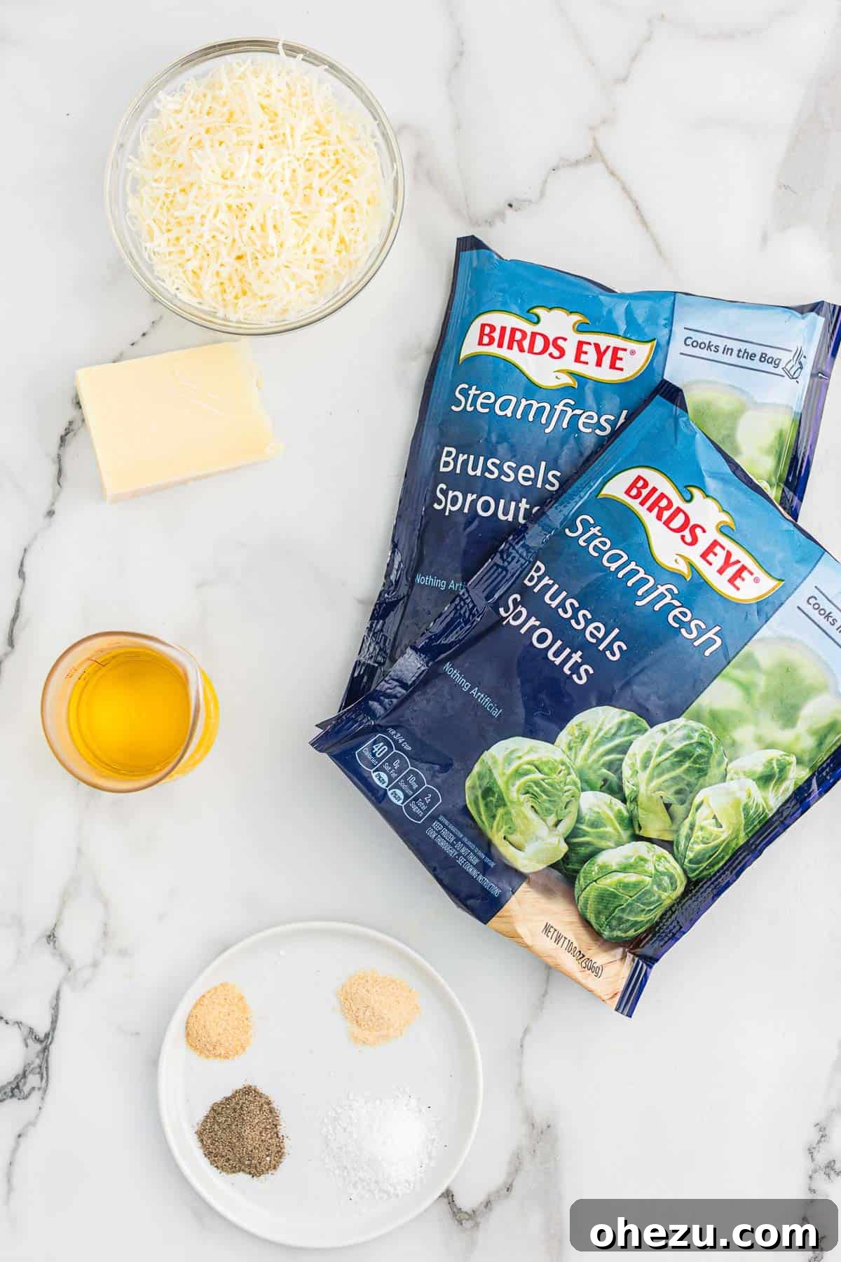 A collection of fresh ingredients laid out on a wooden board, including a bag of frozen Brussels sprouts, a bottle of olive oil, salt, pepper, garlic powder, onion powder, and a bowl of grated Parmesan cheese.