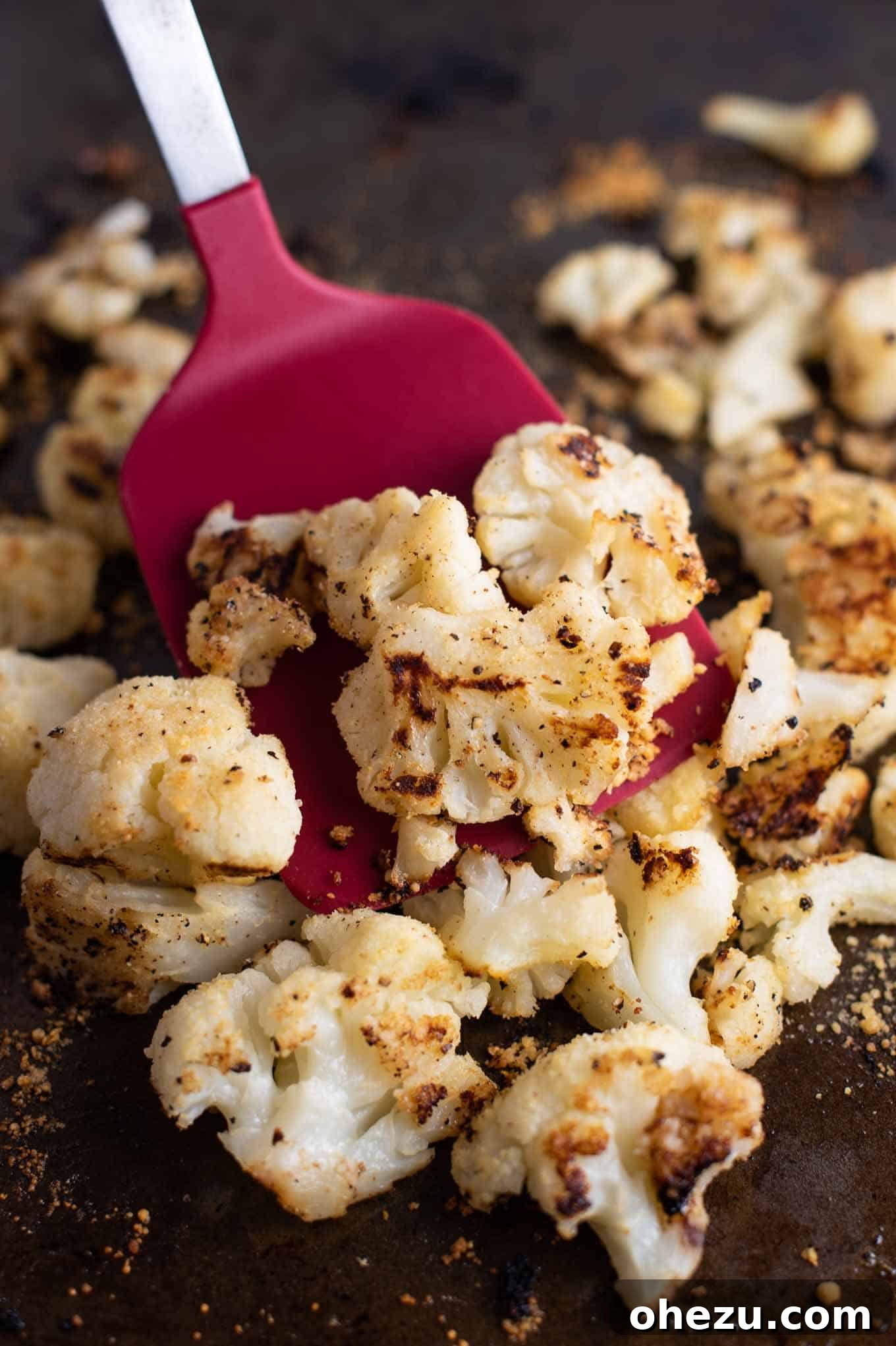 A red spatula scooping up a portion of roasted cauliflower from the baking sheet, highlighting its crispy texture.