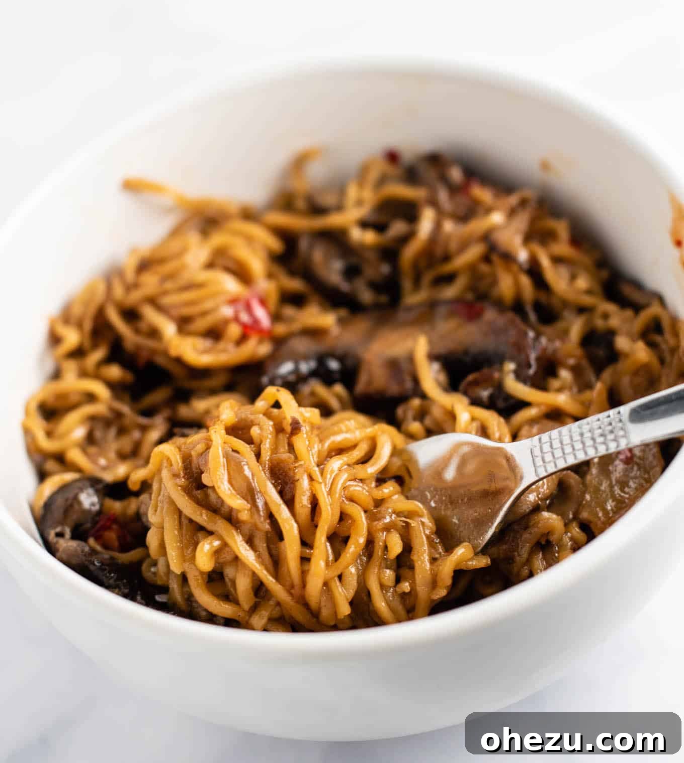ramen noodles twirled on a fork resting on stir fry noodles in a white bowl, enticingly ready to eat