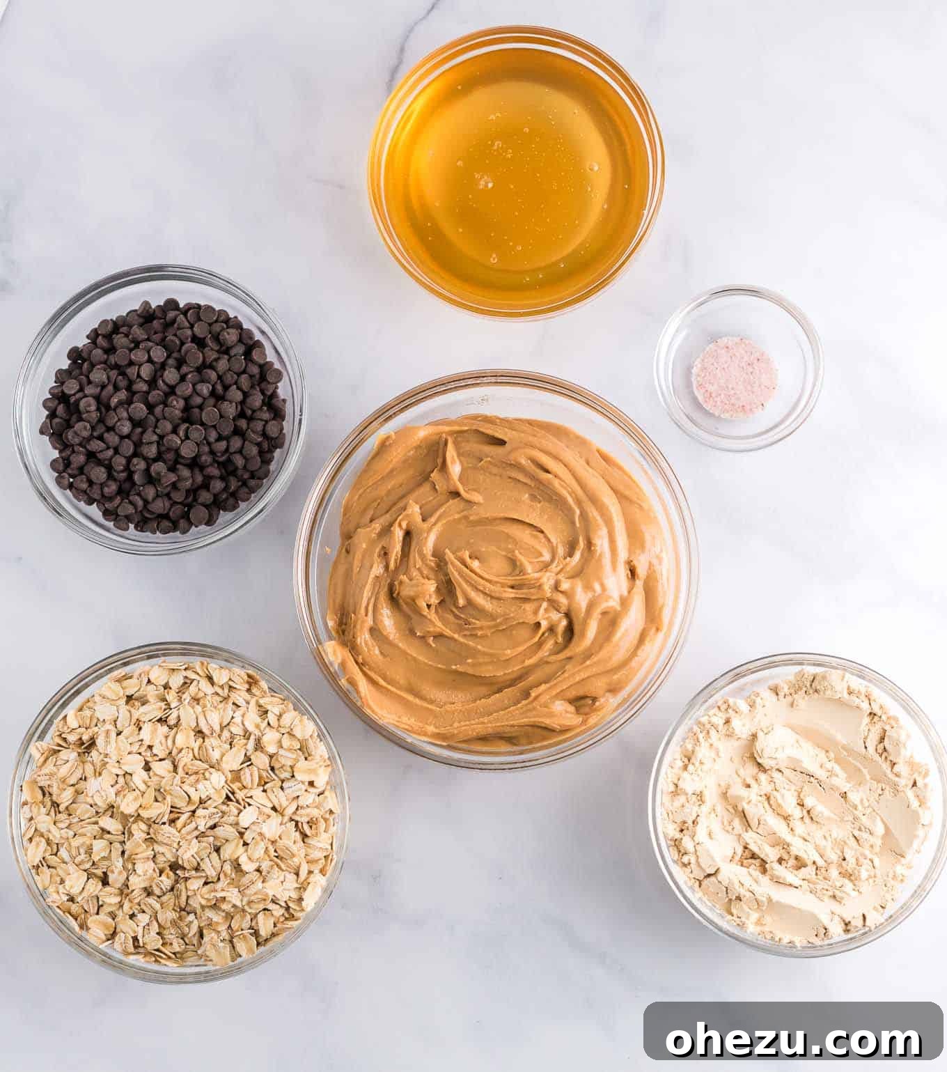 Overhead shot of all six essential ingredients for energy balls: peanut butter, vanilla protein powder, salt, rolled oats, honey, and mini chocolate chips, neatly arranged.