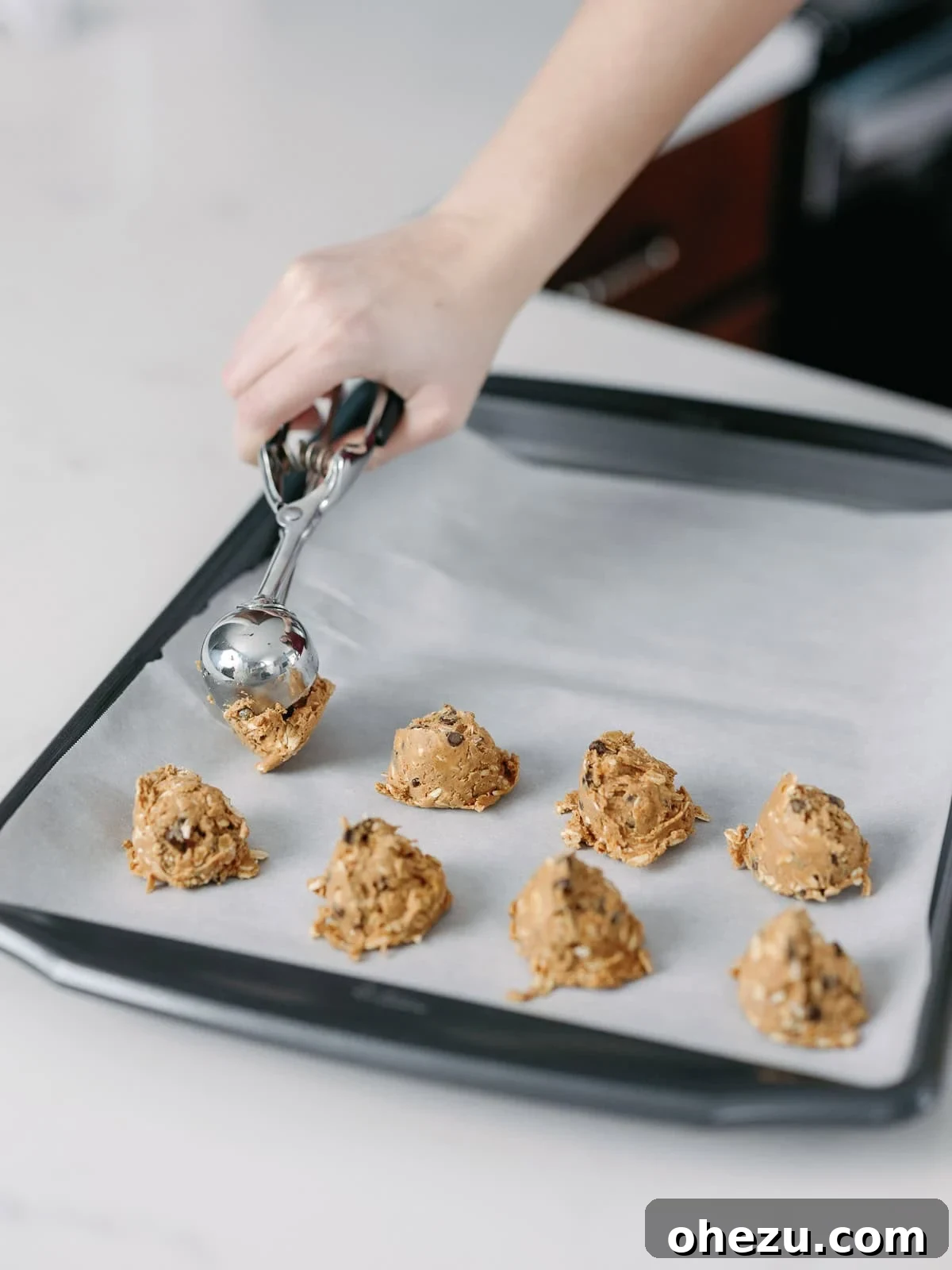 A hand using a mini cookie scoop to portion out energy ball batter onto a parchment-lined baking sheet.