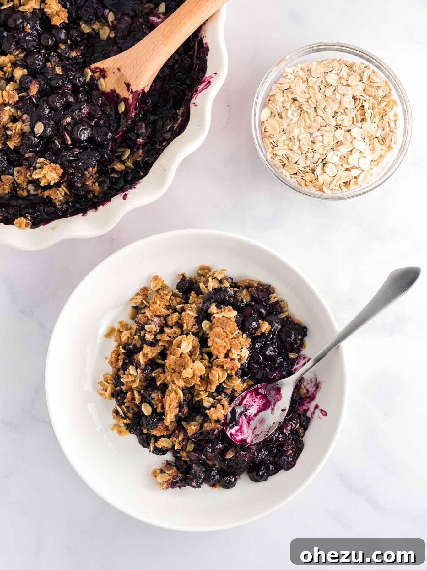 Overhead view of blueberry crisp in a white bowl with a silver spoon, highlighting the rich purple filling and golden topping