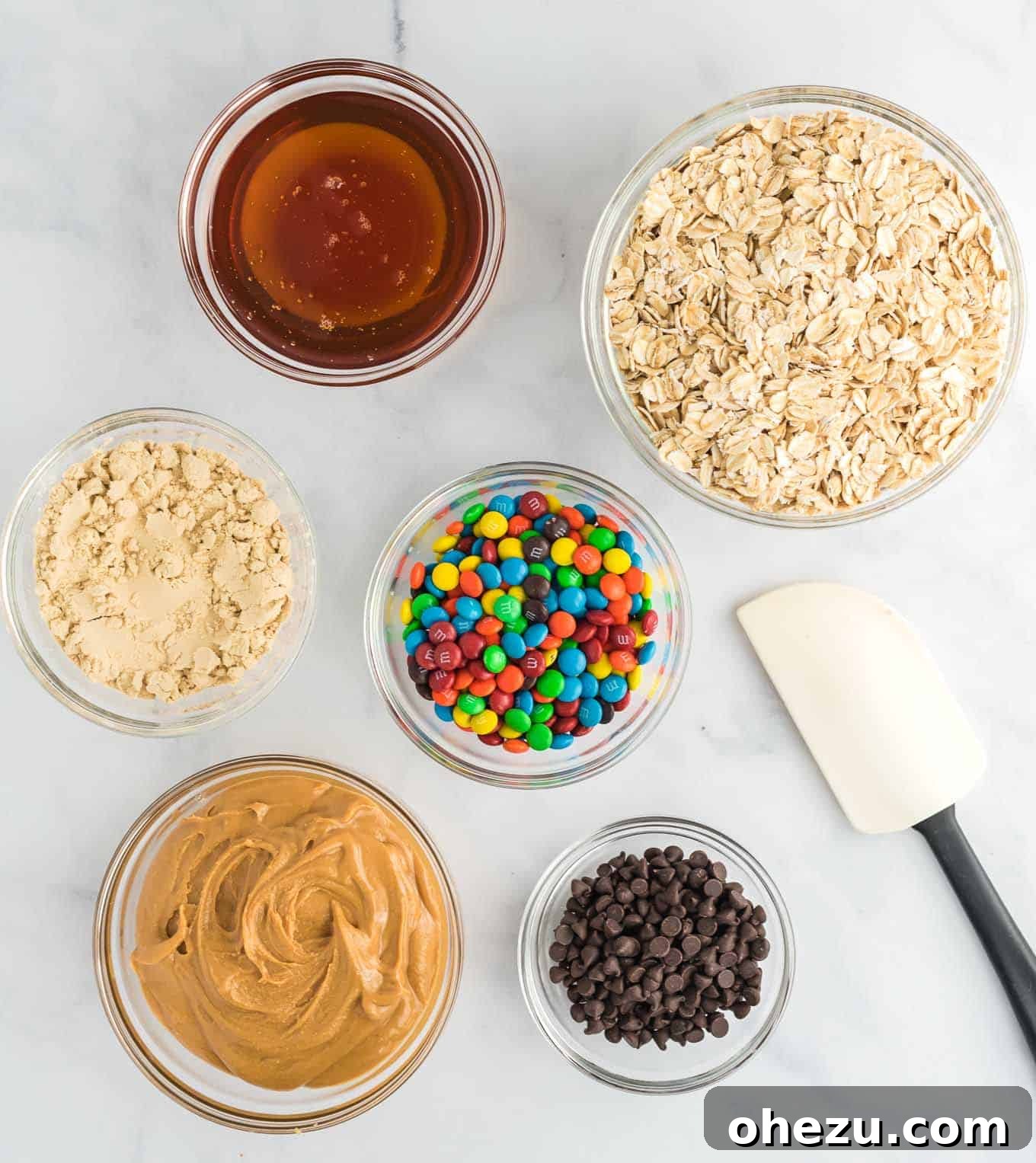 Ultimate Monster Cookie Fuel Bites 4 A selection of raw ingredients for monster cookie energy balls, including peanut butter, honey, protein powder, rolled oats, mini chocolate chips, and mini M&M's, displayed in individual clear bowls.