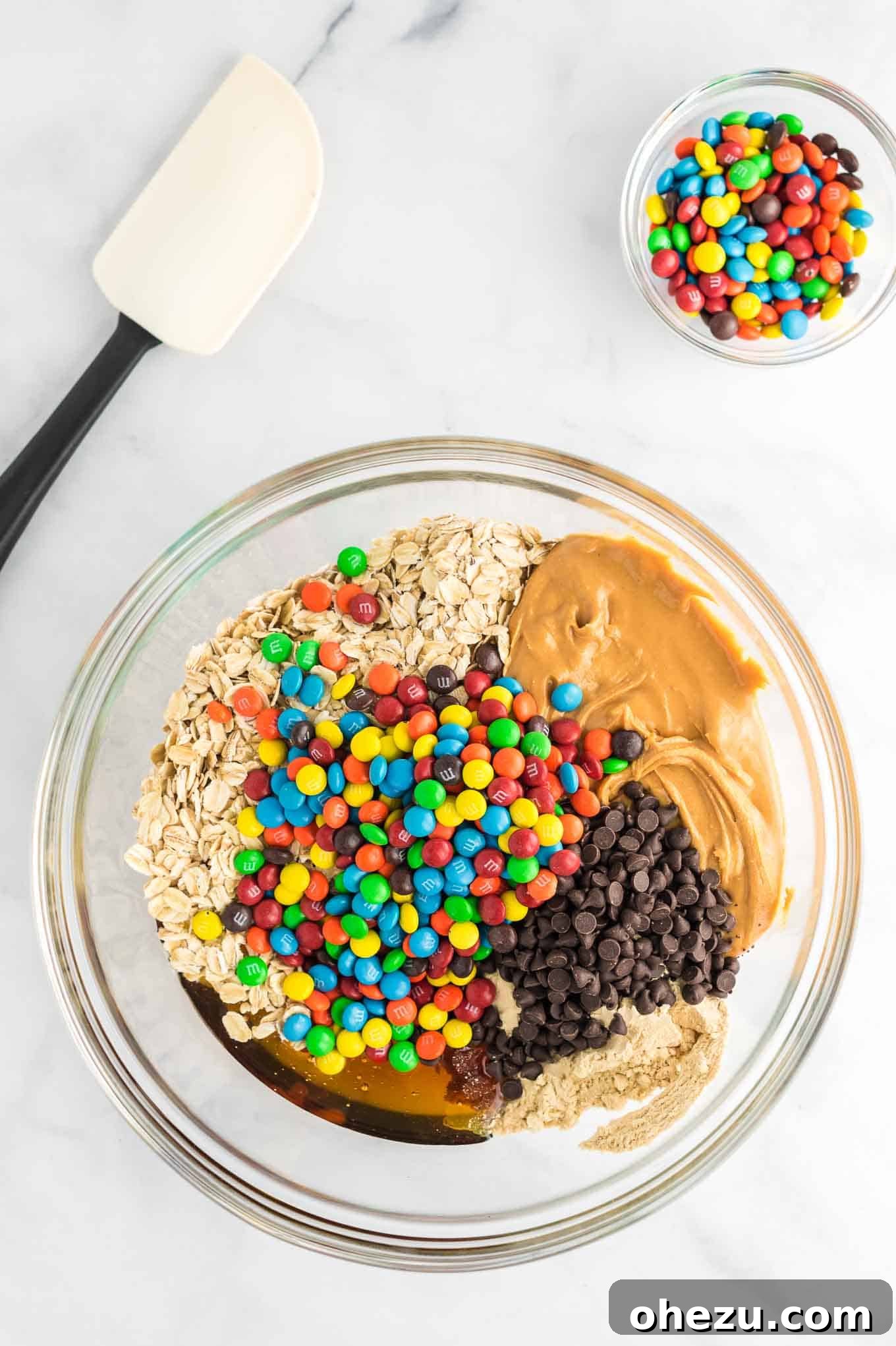 Ultimate Monster Cookie Fuel Bites 5 A large clear mixing bowl filled with all the ingredients for monster cookie energy balls, ready to be mixed: peanut butter, honey, protein powder, rolled oats, mini chocolate chips, and mini M&M's.