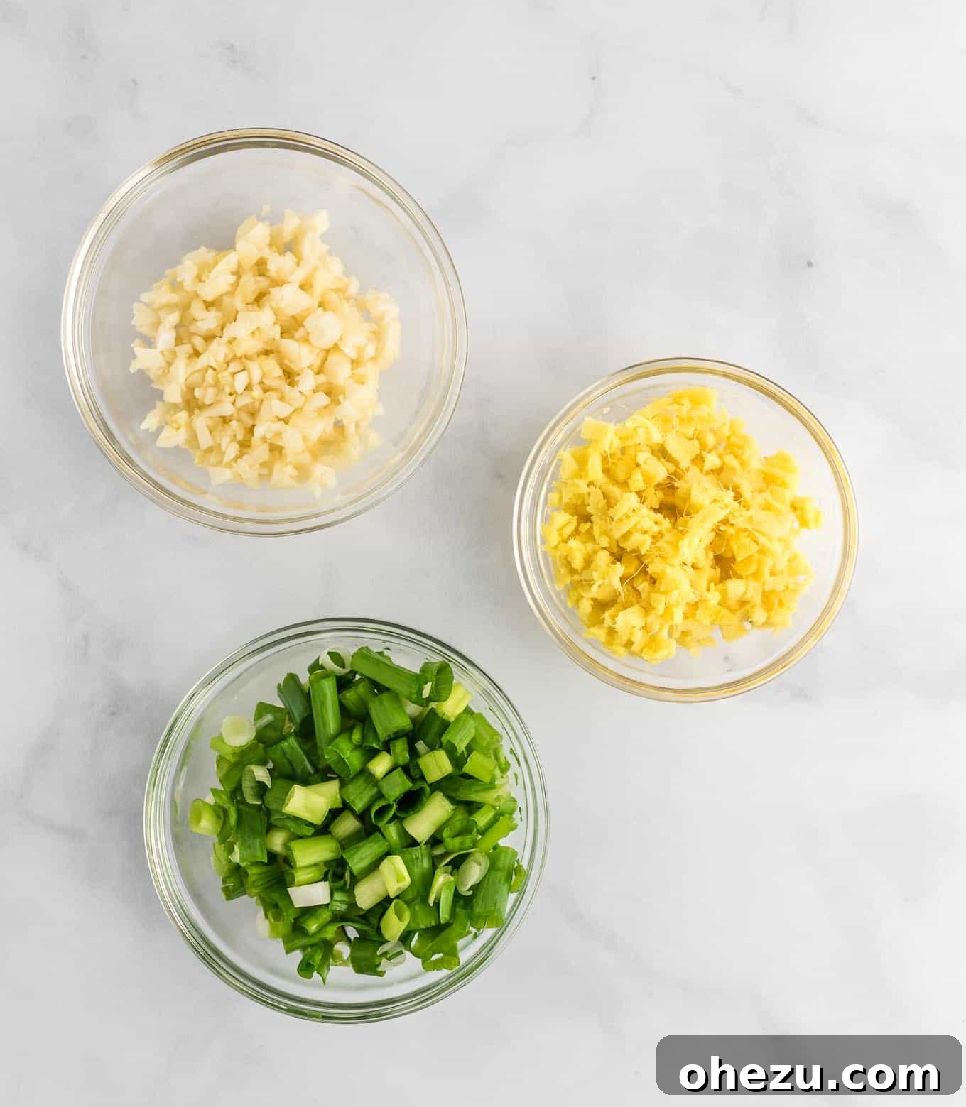 Small clear bowls holding minced green onions, fresh minced ginger, and minced garlic, ready for cooking