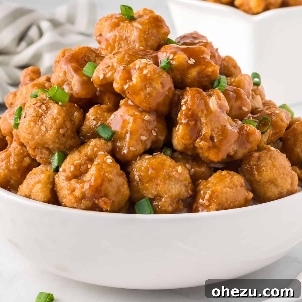 Zesty Orange Glaze 7 A close-up of orange cauliflower pieces in a bowl, generously coated with orange sauce and garnished with fresh green onions and sesame seeds.