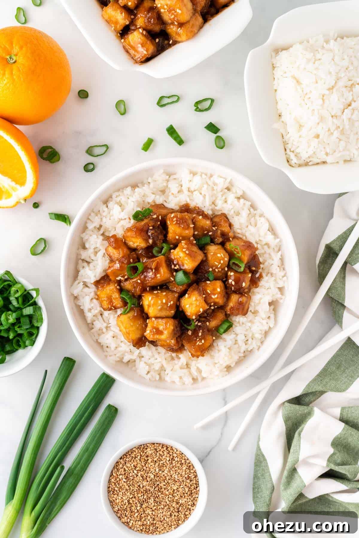 A beautifully composed meal with orange tofu over rice in a bowl, surrounded by its fresh ingredients and garnishes.