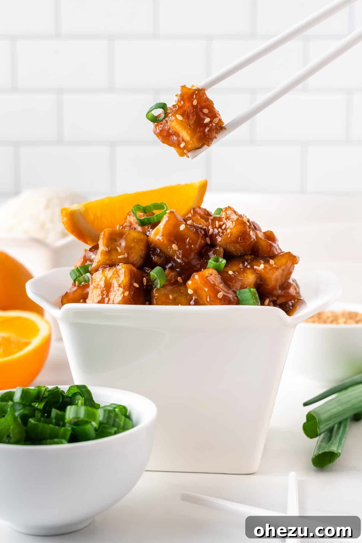 Chopsticks lifting a piece of perfectly glazed orange tofu from a traditional Chinese takeout container, ready for a bite.