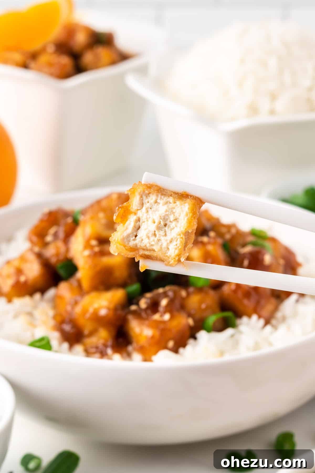 Chopsticks holding a piece of orange tofu with a bite taken out, revealing the crispy texture inside.