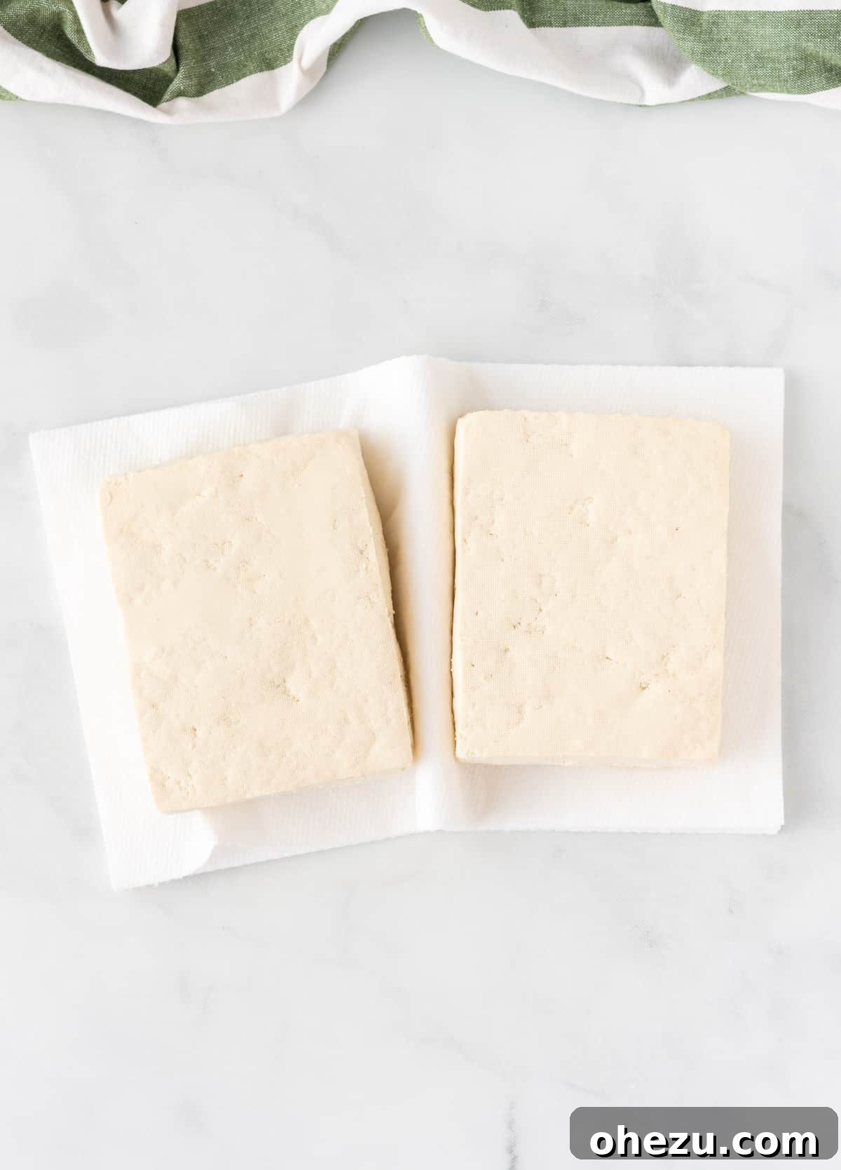 Blocks of extra firm tofu being pressed between two kitchen towels with a heavy object on top to remove moisture.