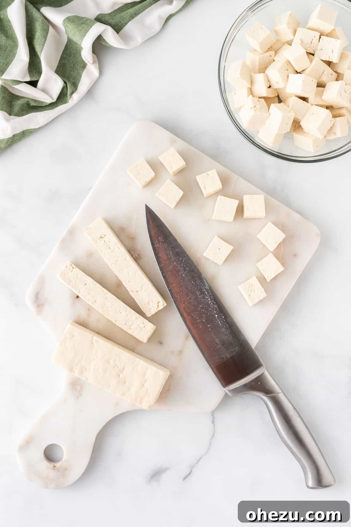 A hand using a knife to cut a block of tofu into perfect cubes on a wooden cutting board.