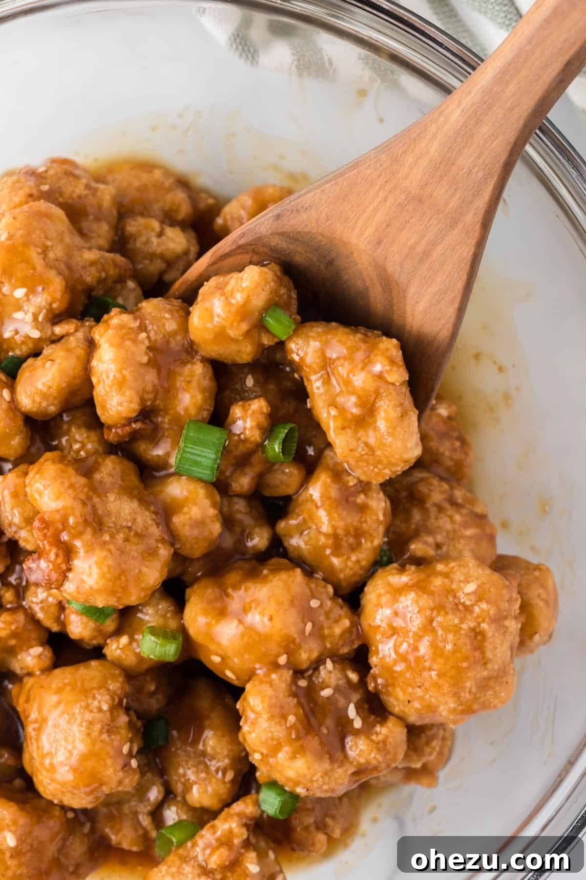 A wooden spoon stirring the orange cauliflower in a large bowl, showing the rich sauce coating the florets