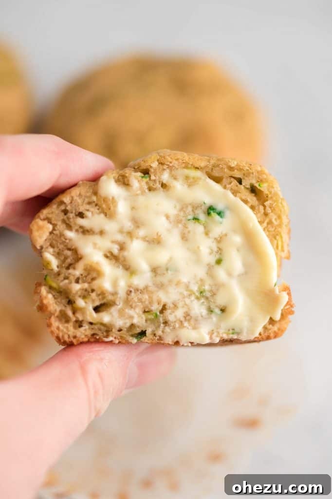 A tantalizing close-up of a zucchini muffin, perfectly split and adorned with melting butter, inviting a delicious bite, with more muffins in the background.