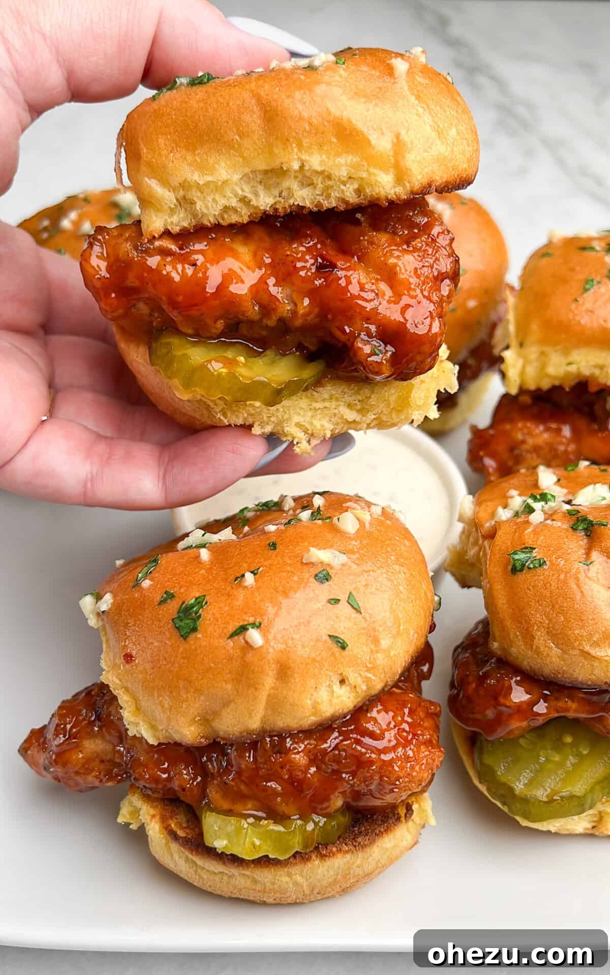 A close-up view of two freshly prepared Hot Honey Chicken Sliders on a rustic white plate, highlighting the golden-brown crispy chicken, glossy hot honey glaze, and vibrant green pickles, with a small bowl of ranch in the background.