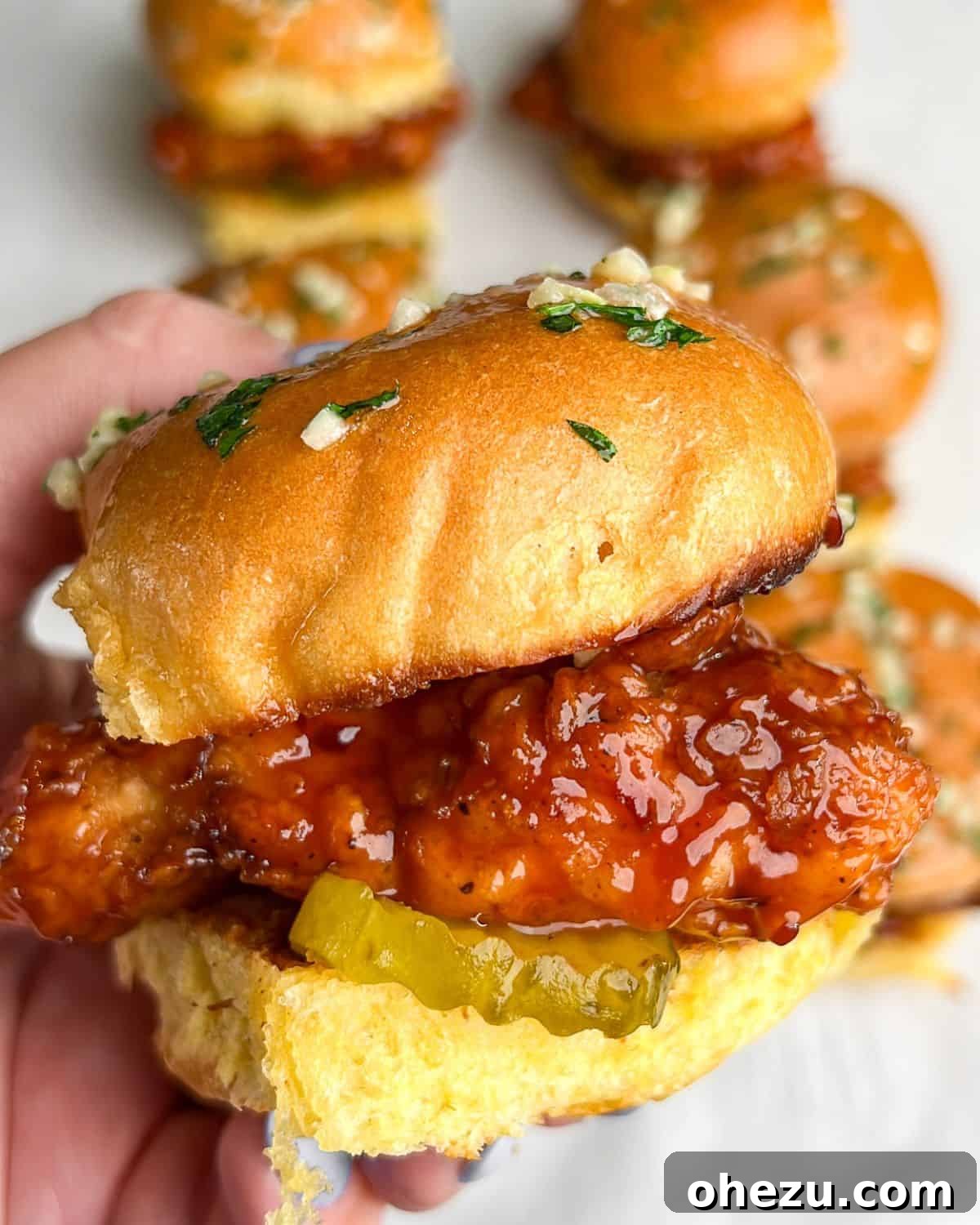 A hand holding a delicious Hot Honey Chicken Slider, highlighting the crispy chicken, hot honey glaze, and visible pickles within the brioche bun.