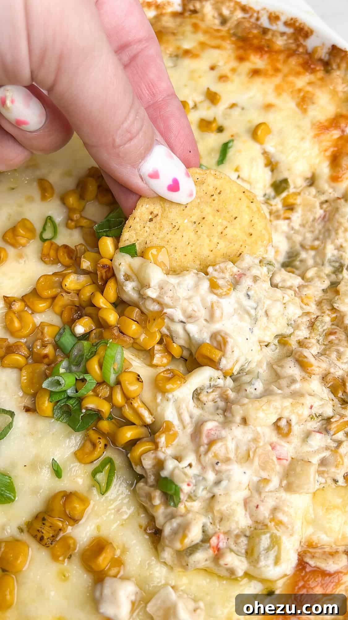 A hand reaching into a casserole dish of hot corn dip with a tortilla chip, showing the dip's appealing texture and readiness for enjoyment.