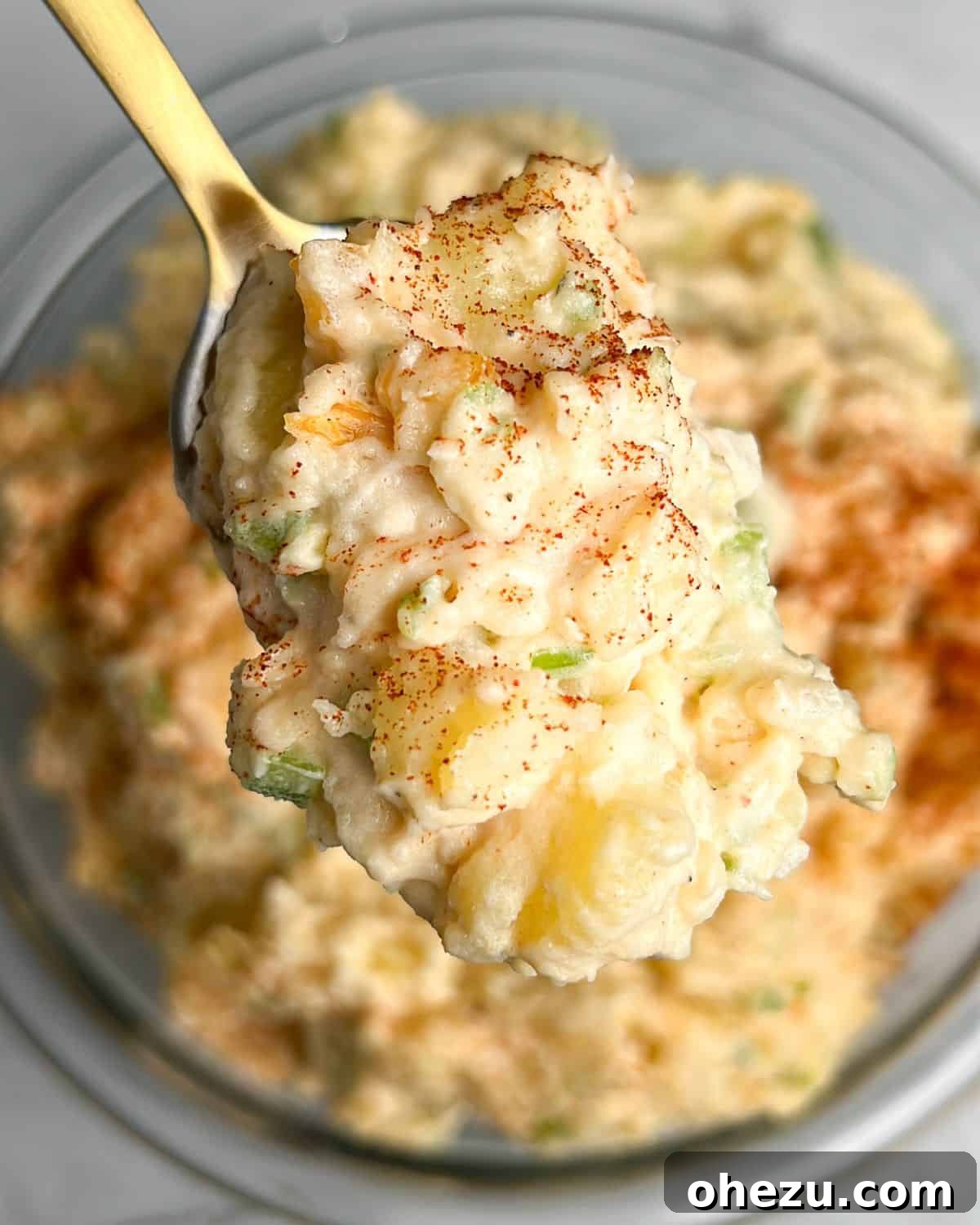 A serving spoon lifting a spoonful of eggless potato salad from a large bowl, showcasing its creamy texture and visible vegetable pieces.